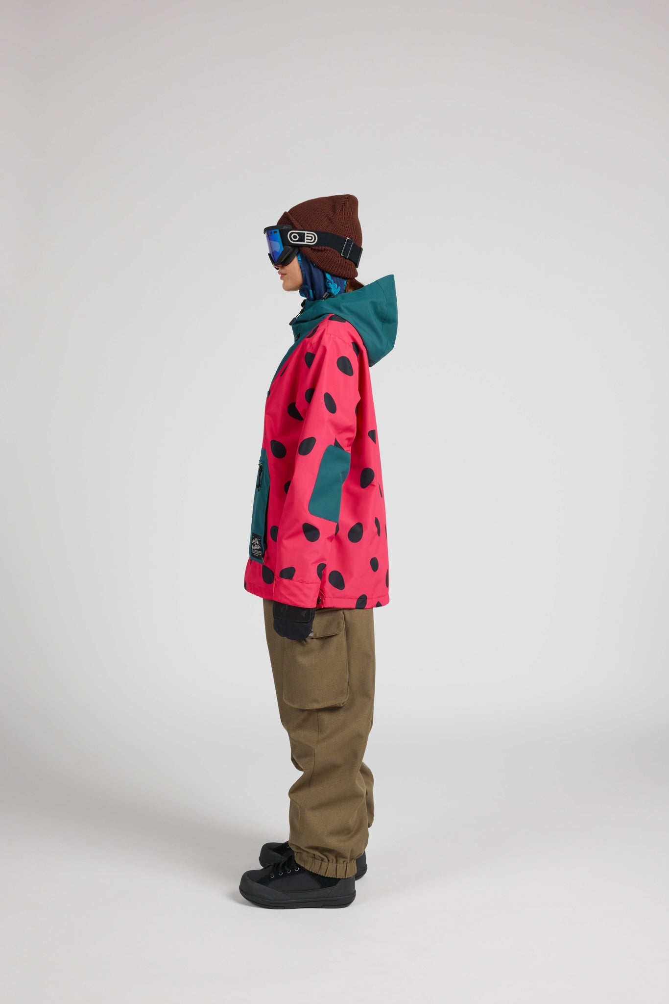 A model shown in profile wearing a vibrant pink ski jacket with black spots, a teal hood, brown pants, and a brown beanie against a neutral gray studio background.