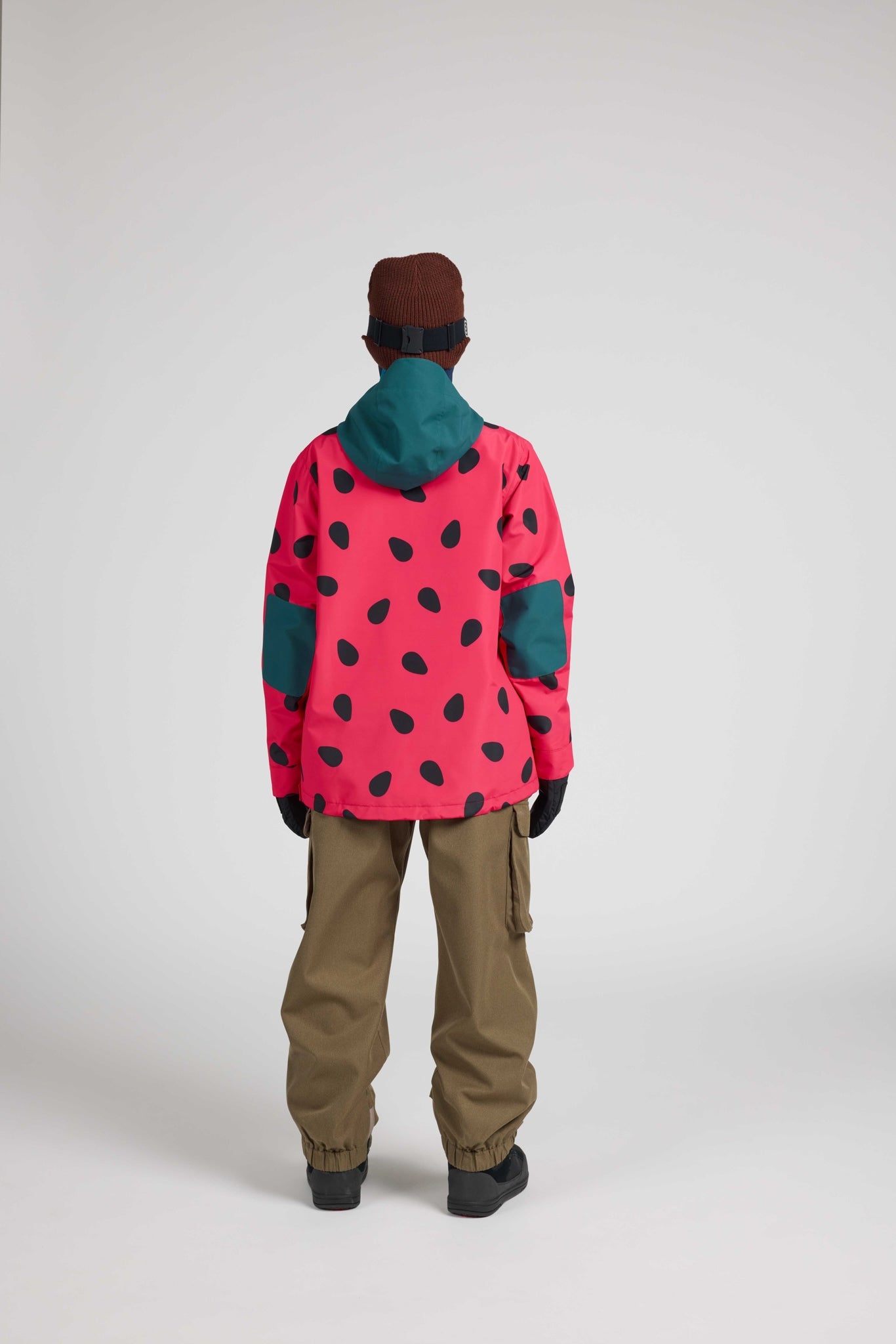 A model shown from the back wearing a vibrant pink ski jacket with a black spotted pattern, teal hood and elbow patches, olive cargo pants, and a brown beanie against a light gray studio background.