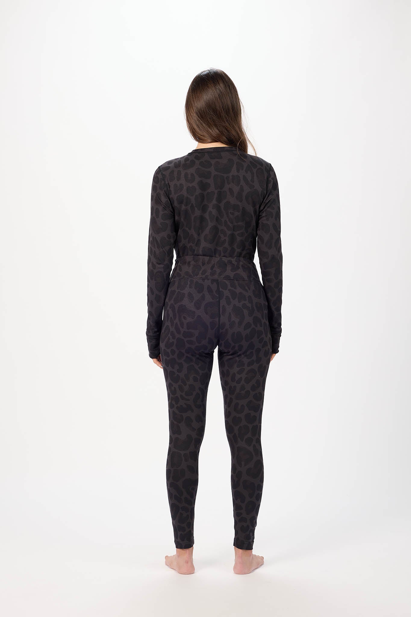 A woman viewed from behind wearing a long-sleeve one-piece jumpsuit with a dark gray and black leopard print, standing against a plain white studio background.