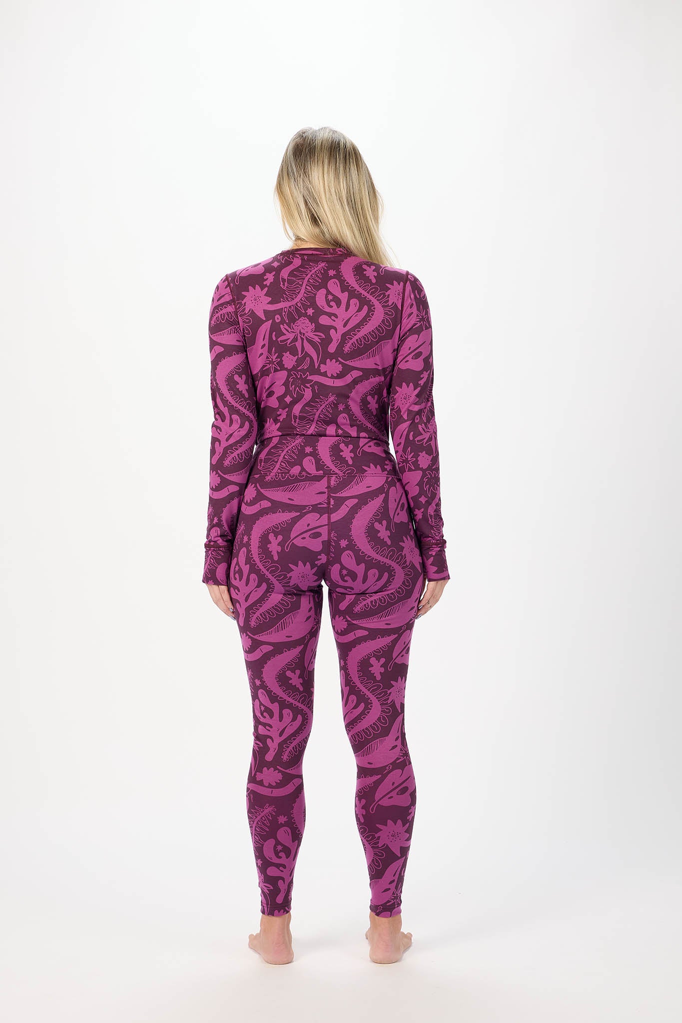 A woman stands barefoot against a plain white studio background wearing a long-sleeve base layers with an allover abstract floral or paisley-like pattern in two shades of purple. viewed from behind