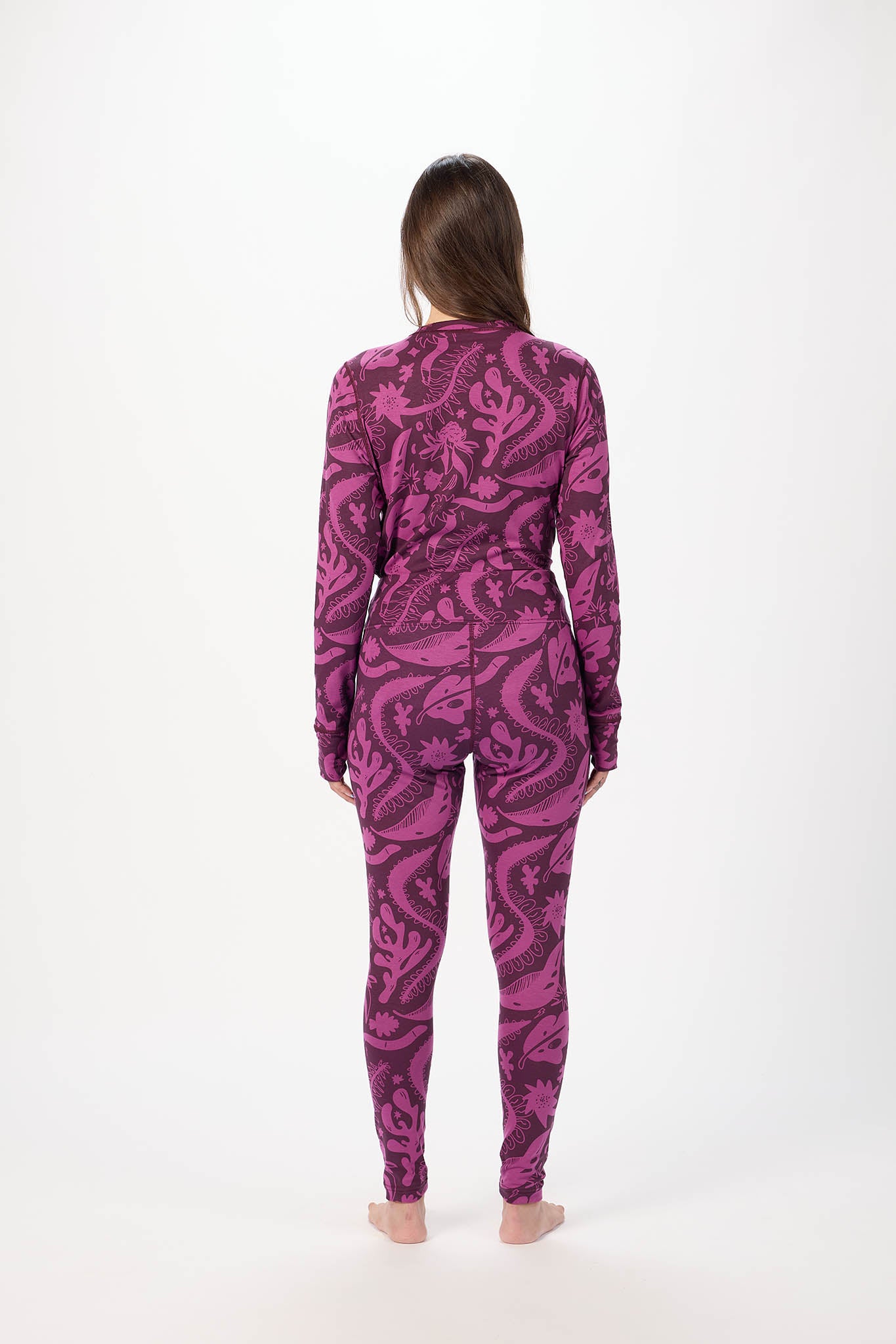A woman viewed from behind wearing a matching long-sleeve top and leggings set with a dark purple and magenta paisley pattern, standing against a plain white studio background.
