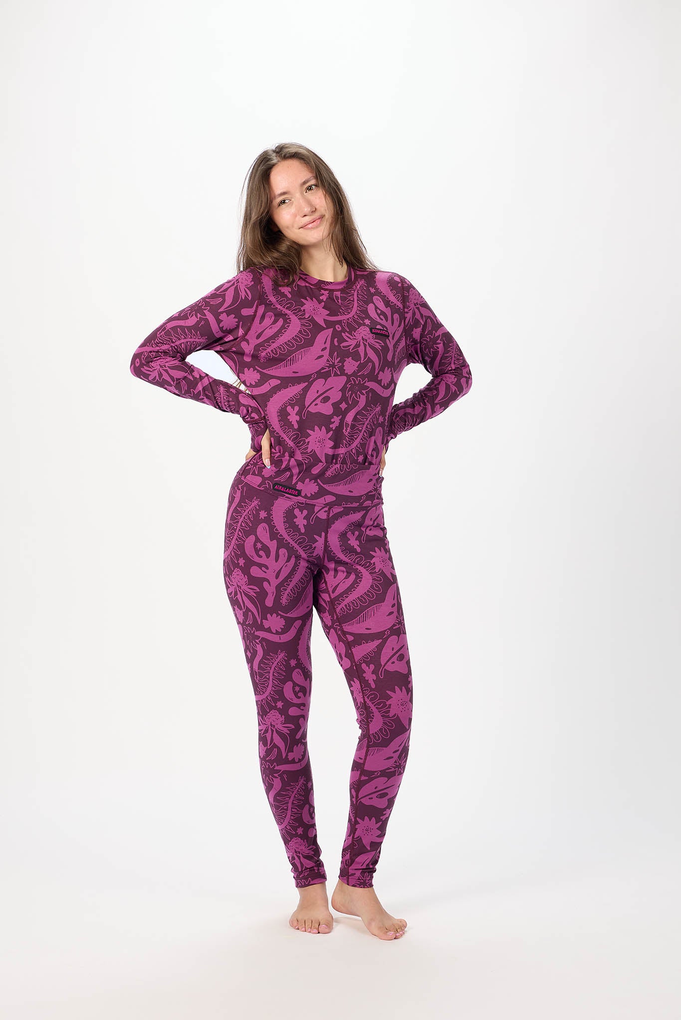 A woman modeling a matching two-piece long-sleeve top and leggings set with a dark purple and magenta paisley pattern, standing against a plain white studio background.