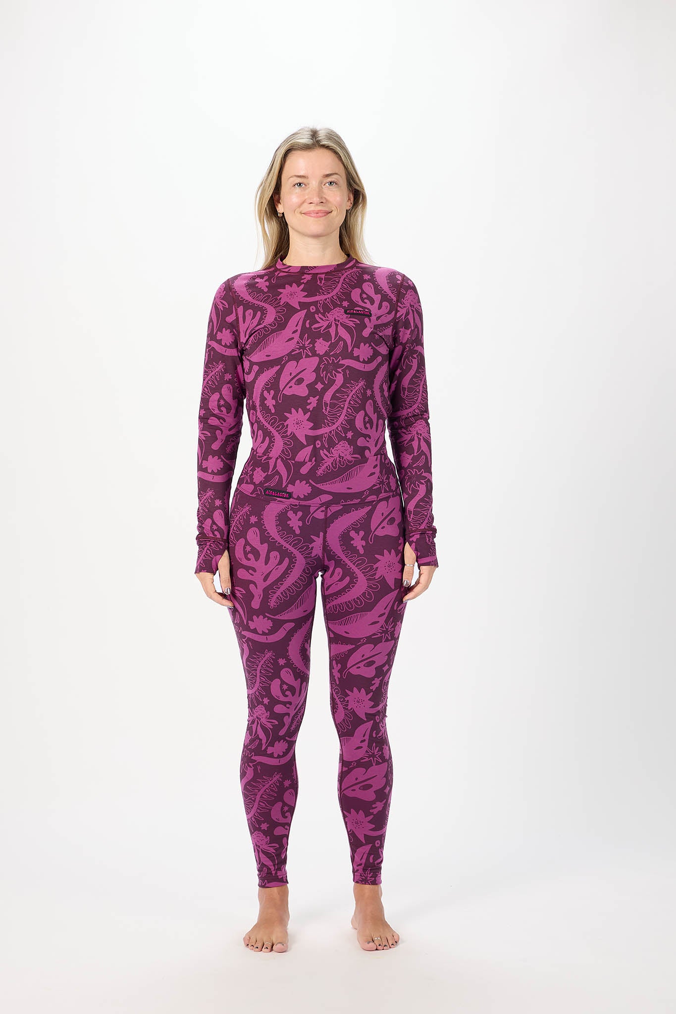 A woman stands barefoot against a plain white studio background wearing a long-sleeve base layers with an allover abstract floral or paisley-like pattern in two shades of purple.