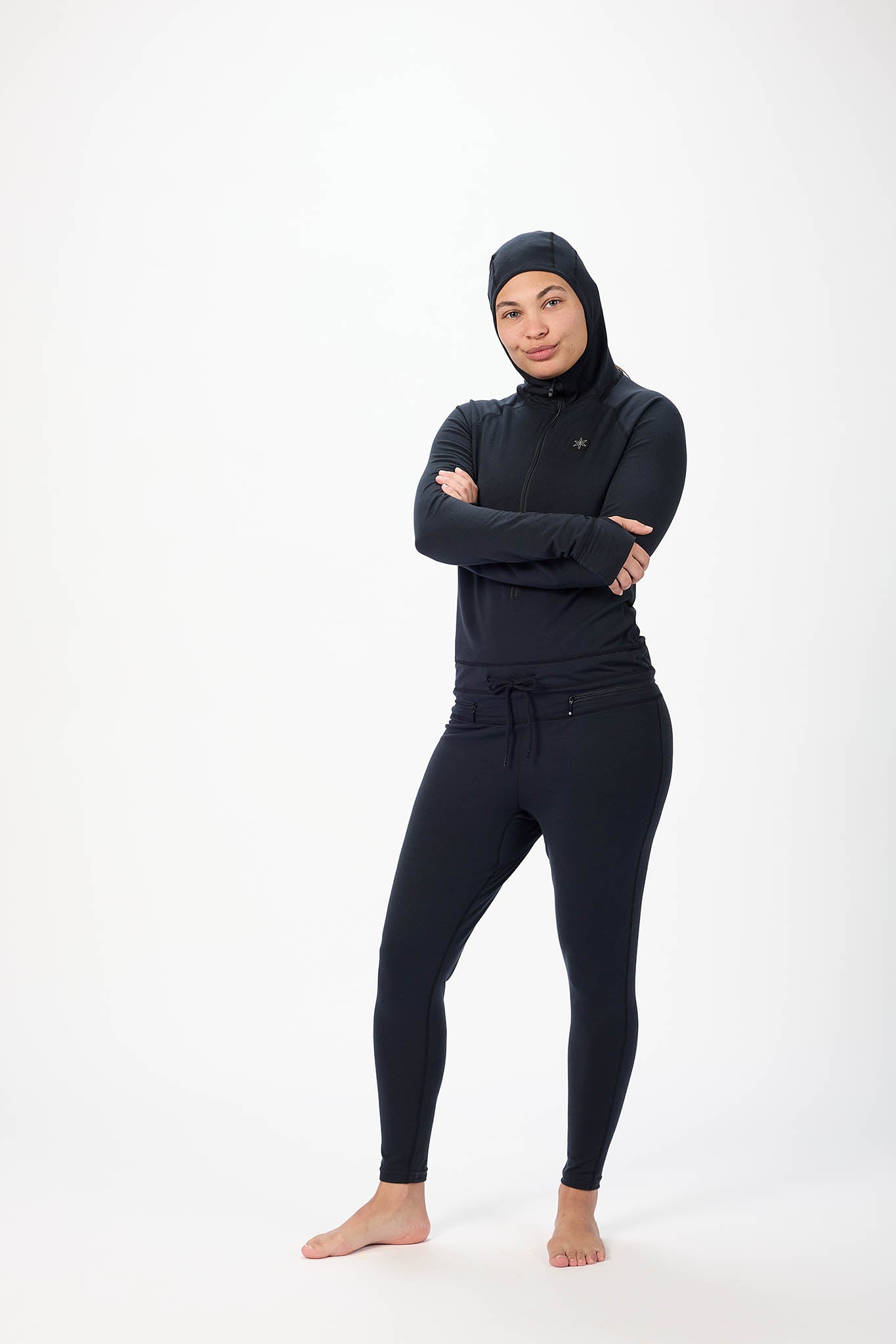 A woman wearing a black full-body athletic suit with a high-neck zip-front top, matching pants, and a hood, standing against a plain white studio background.