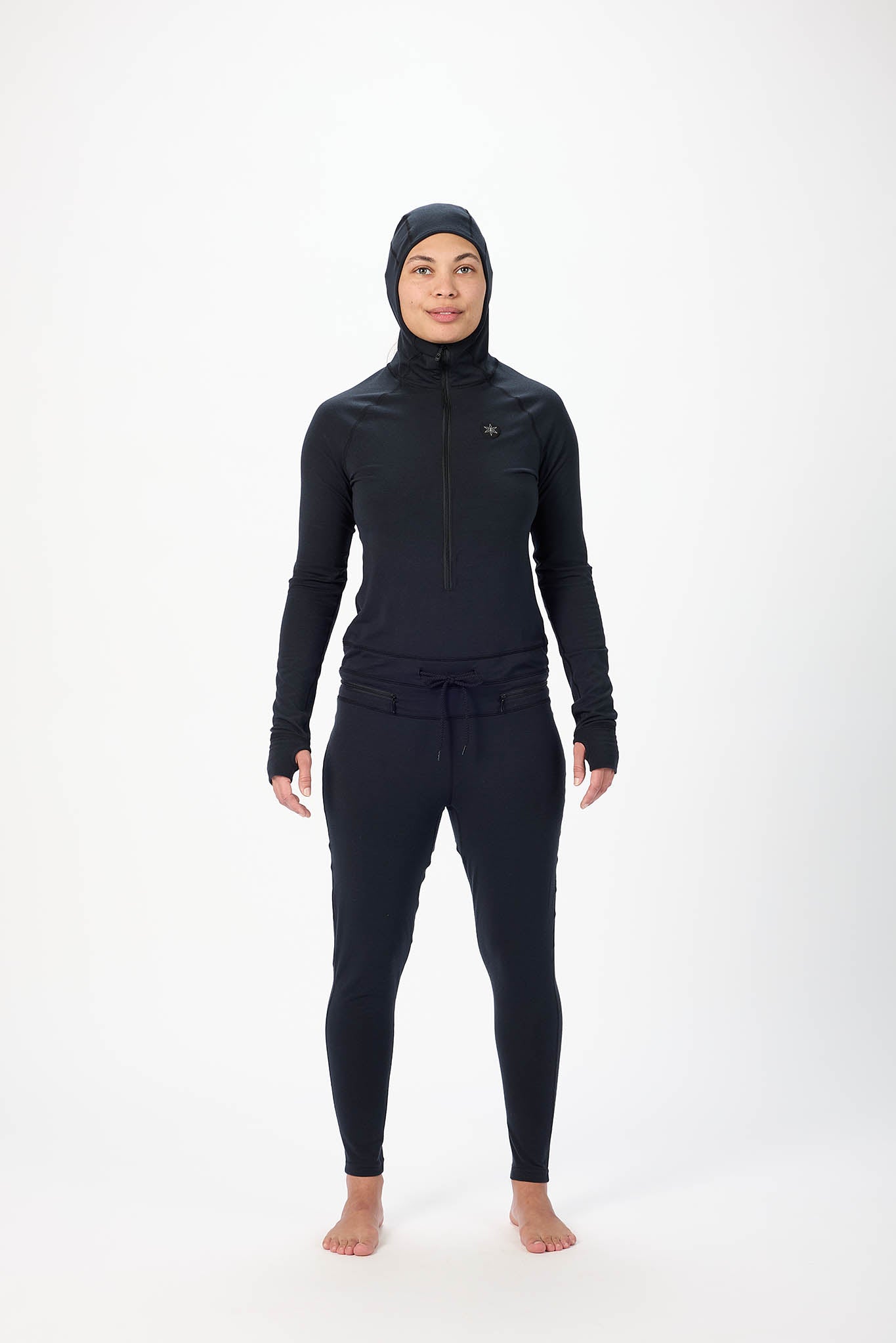 A woman wearing a black full-body athletic suit with a high-neck zip-front top, matching pants, and a hood, standing against a plain white studio background.