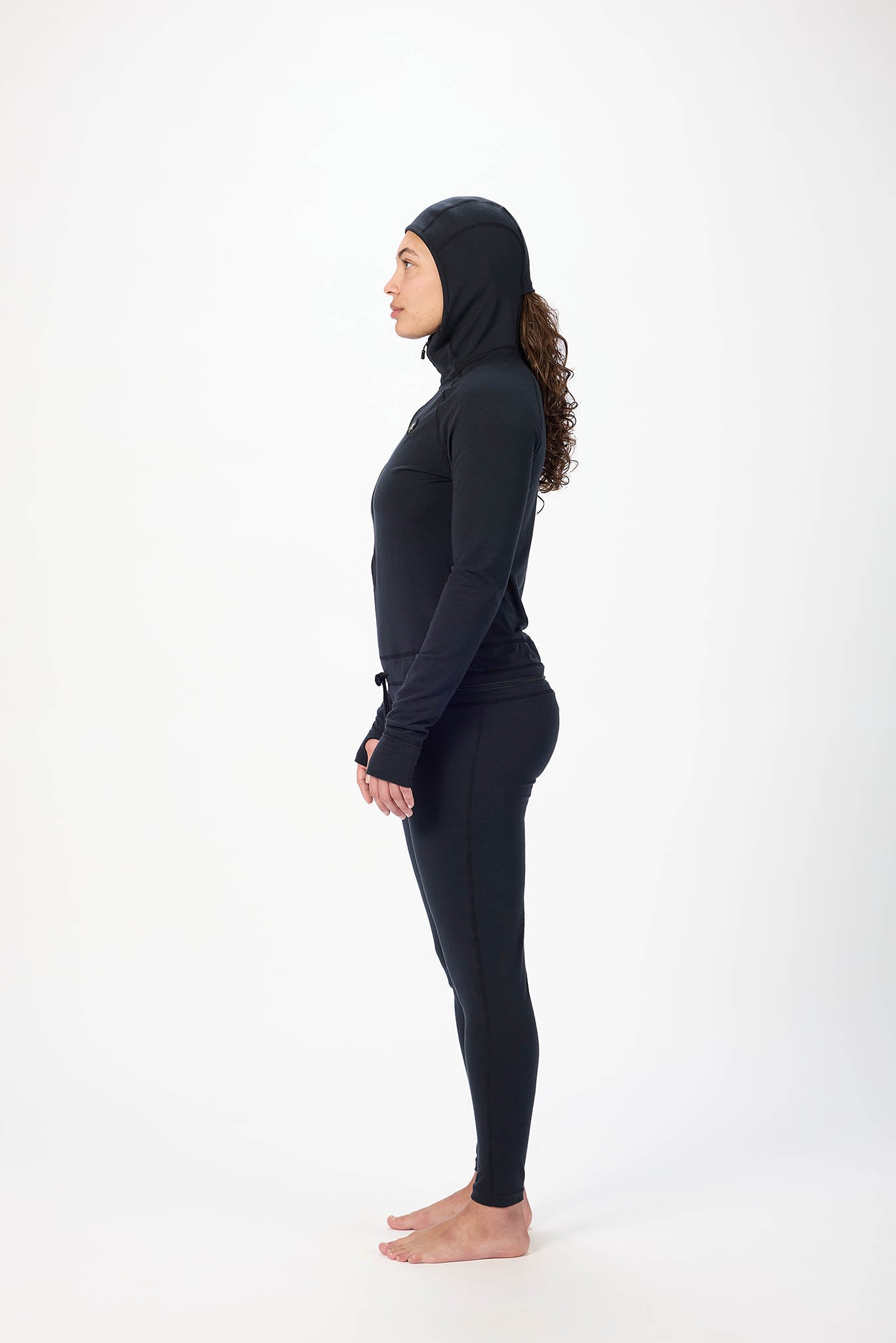 in side profile A woman wearing a black full-body athletic suit with a high-neck zip-front top, matching pants, and a hood, standing against a plain white studio background.