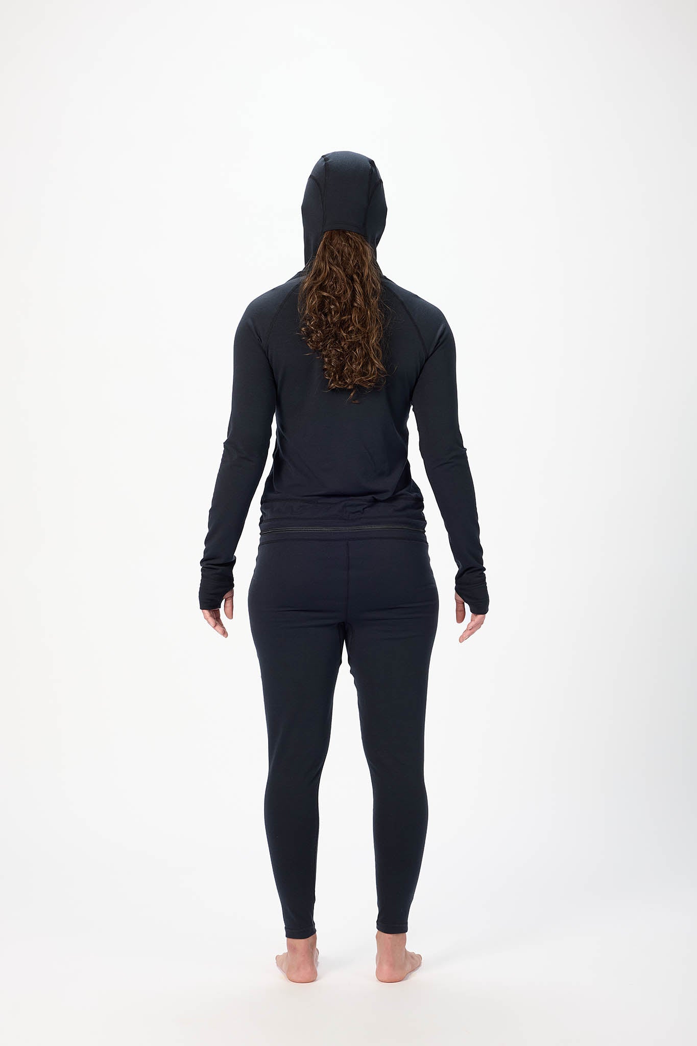 A woman viewed from behind wearing a matching black long-sleeve hooded top and leggings set, standing barefoot against a plain white studio background.
