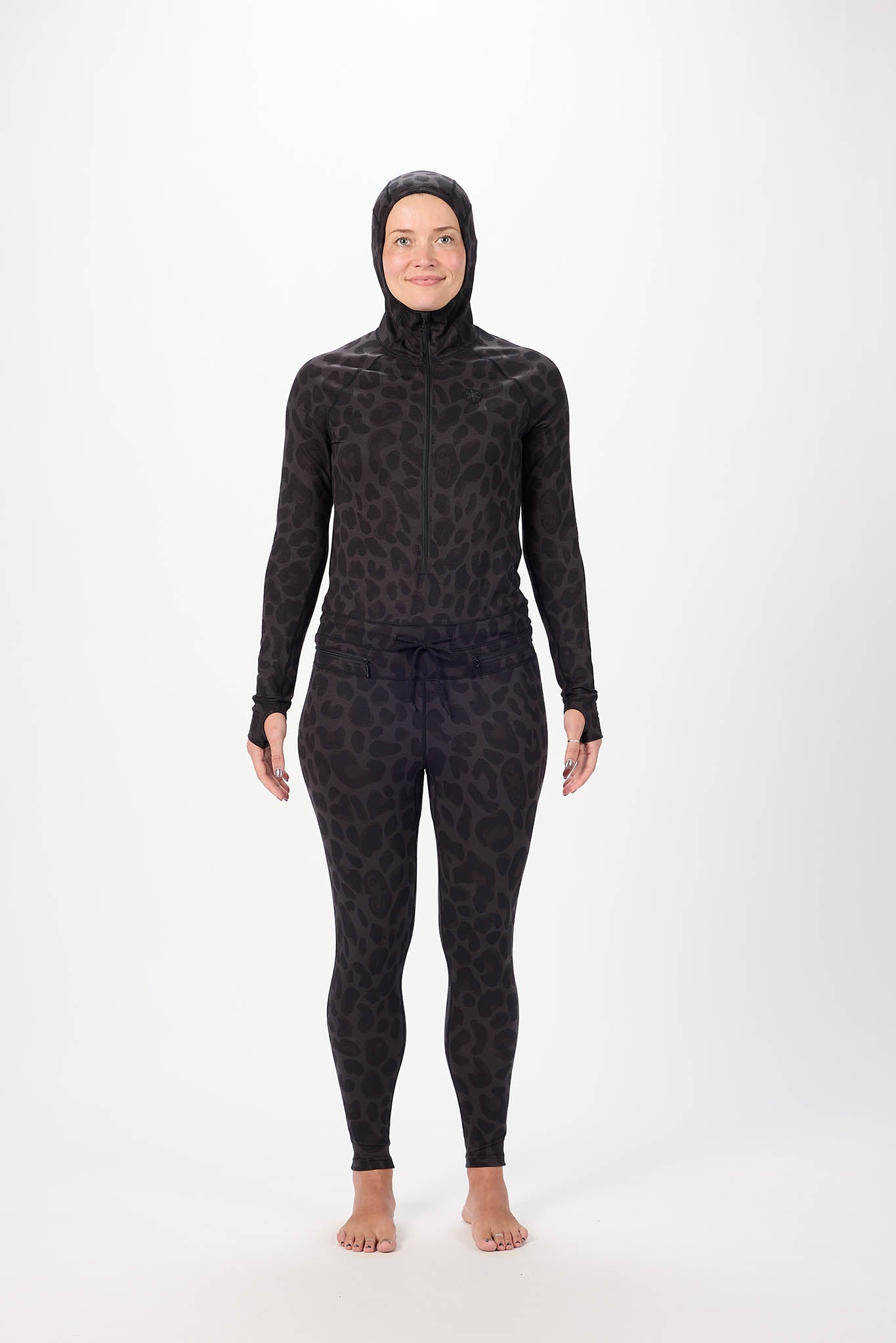 A woman wearing a full-body black leopard-print onesie with a hood and face mask, standing against a plain white studio background.
