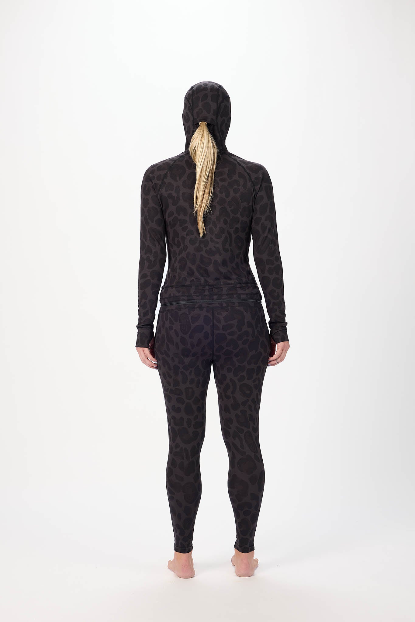 A woman wearing a full-body black leopard-print onesie with a hood and face mask, standing against a plain white studio background viewed from behind.