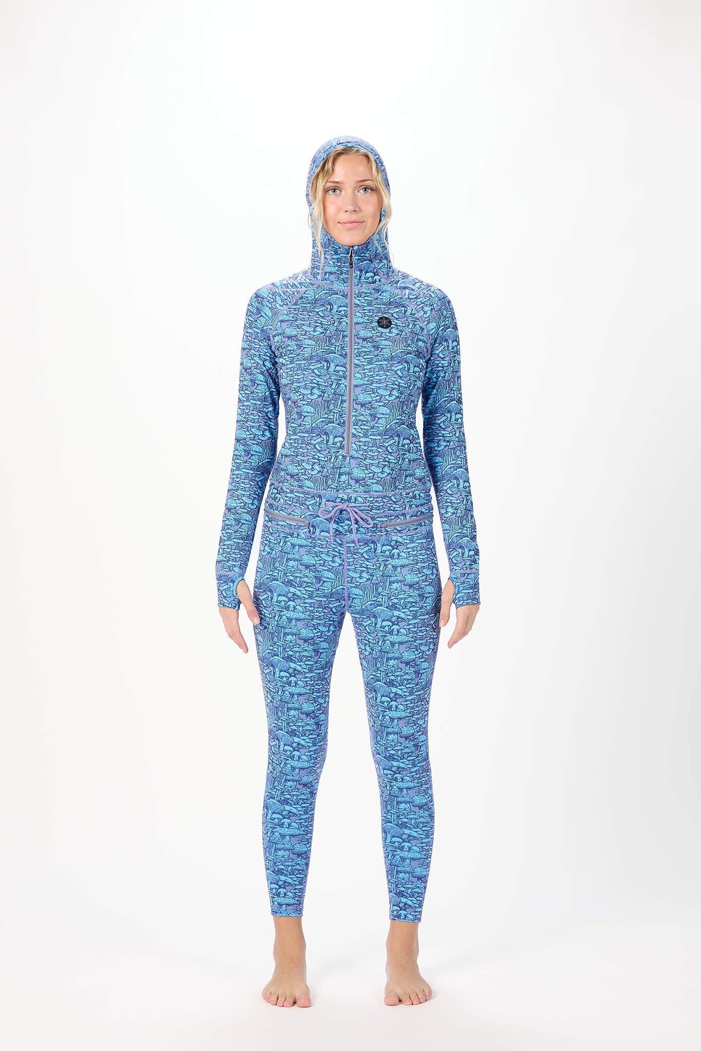 A woman modeling a full-body hooded one-piece with a light white background and a repeating print of Mushrooms standing against a plain white studio background.