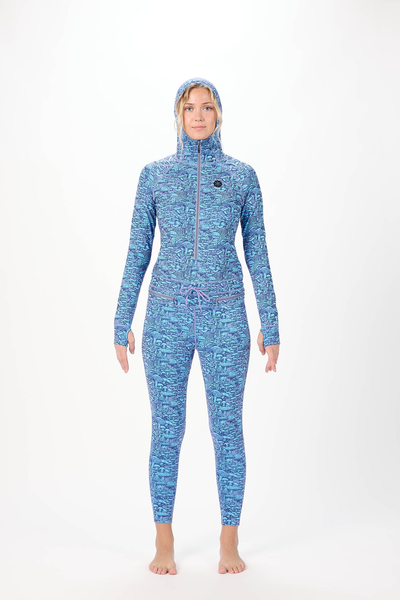 A woman modeling a full-body hooded one-piece with a light white background and a repeating print of Mushrooms standing against a plain white studio background.