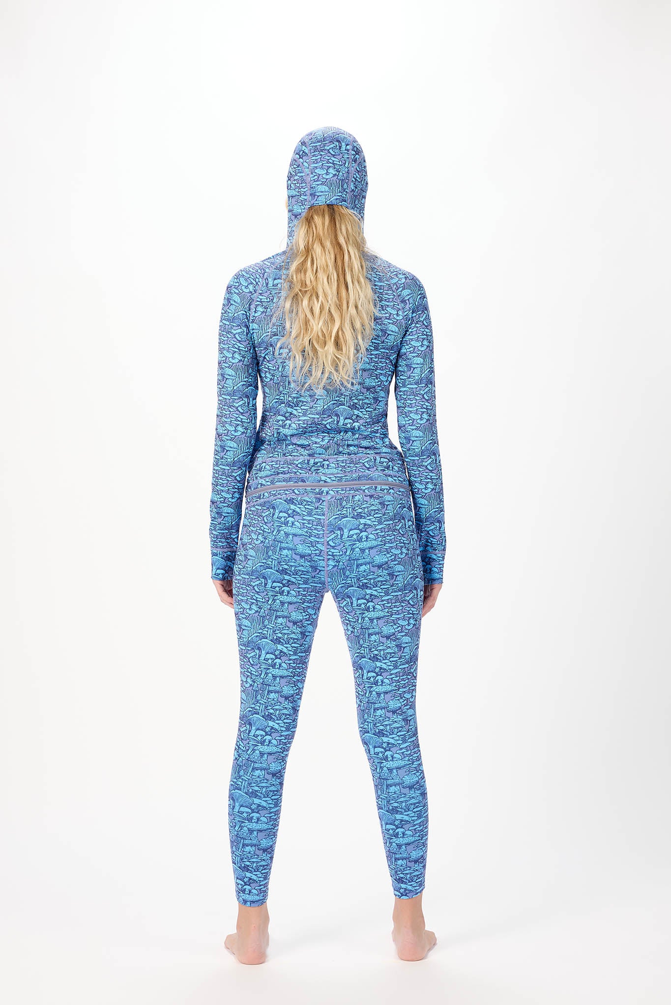 A woman modeling a full-body hooded one-piece with a light white background and a repeating print of Mushrooms standing against a plain white studio background.