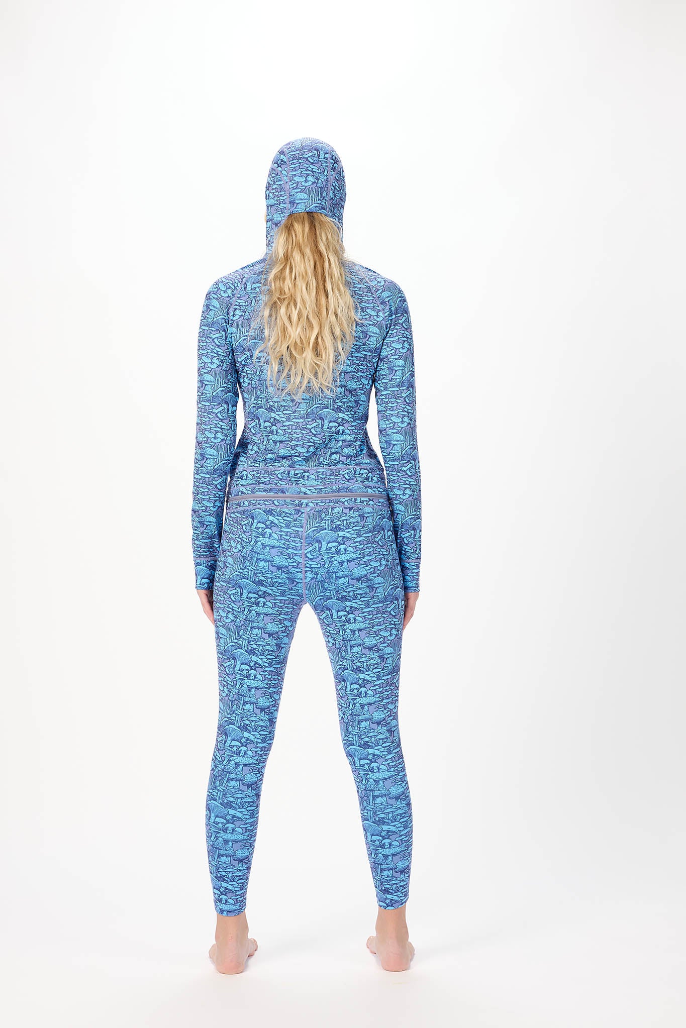 A woman modeling a full-body hooded one-piece with a light white background and a repeating print of Mushrooms standing against a plain white studio background.