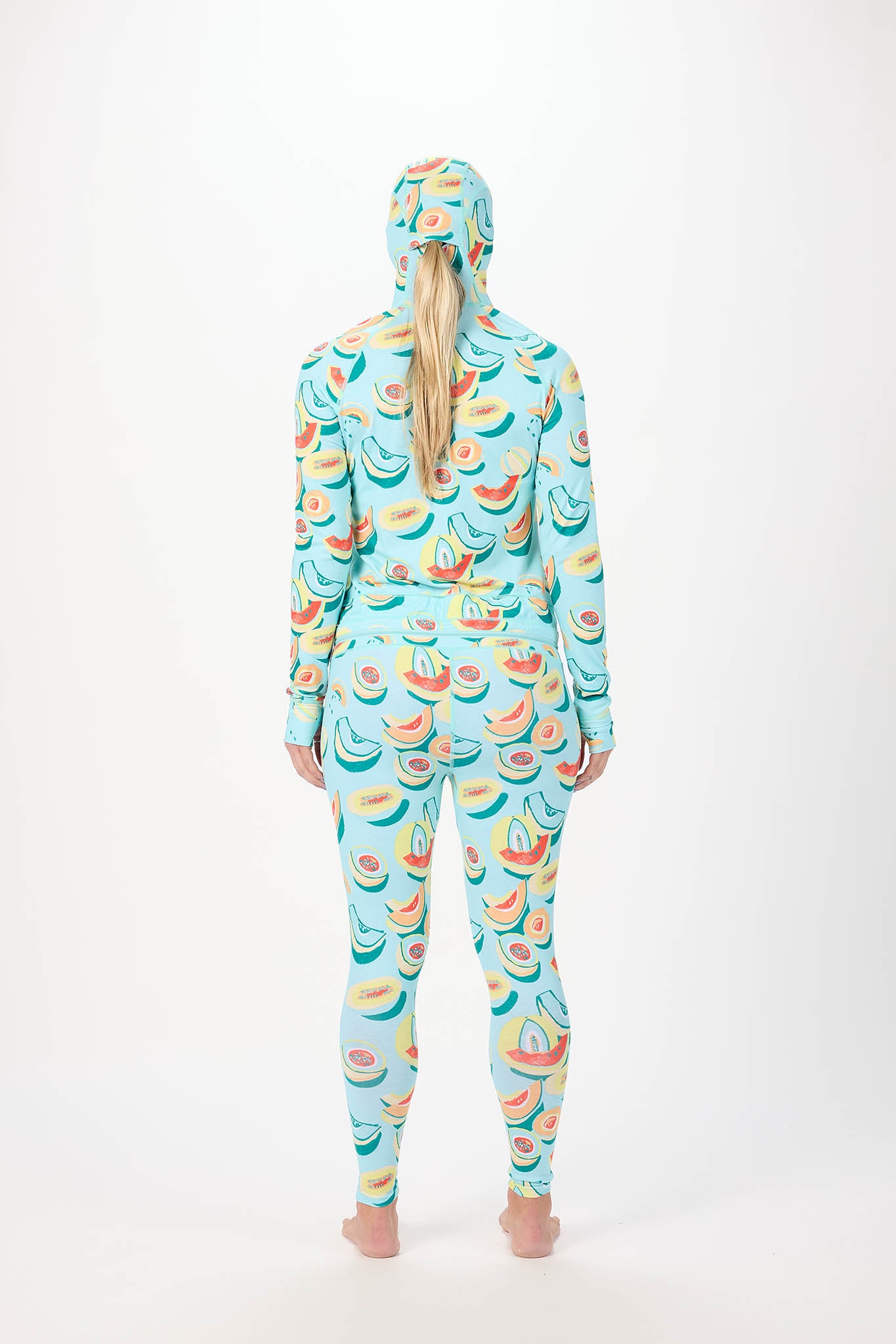 A person viewed from the back wearing a hooded long-sleeve one-piece jumpsuit covered in a colorful avocado and watermelon print on a light blue base, standing against a plain white studio background.