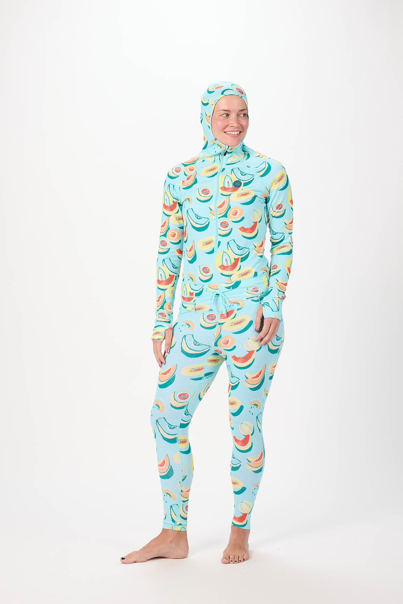 A woman modeling a full-body hooded one-piece with a light white background and a repeating print of bananas, watermelons, and strawberries, standing against a plain white studio background.