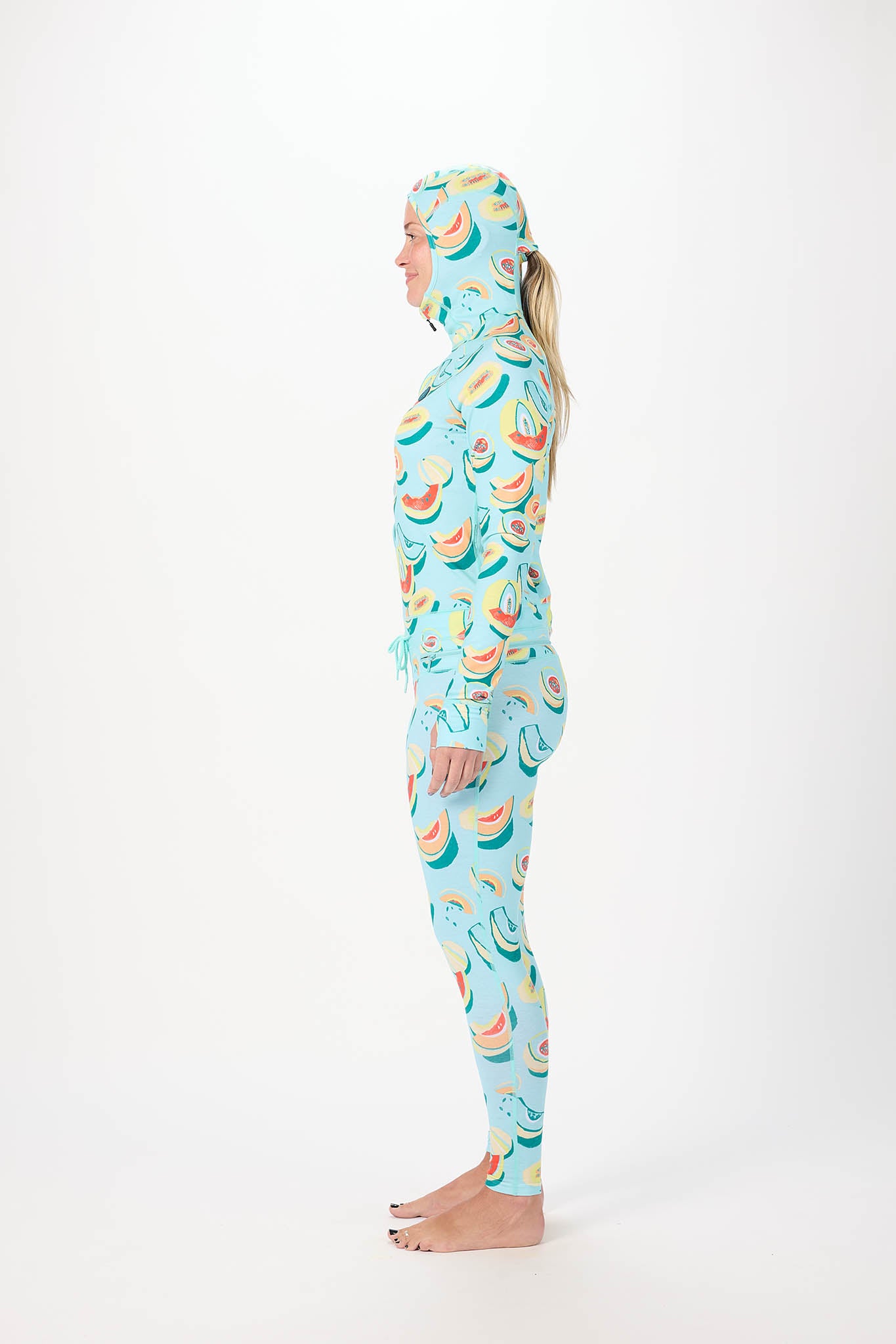 in side profile A woman modeling a full-body hooded one-piece with a light white background and a repeating print of bananas, watermelons, and strawberries, standing against a plain white studio background.