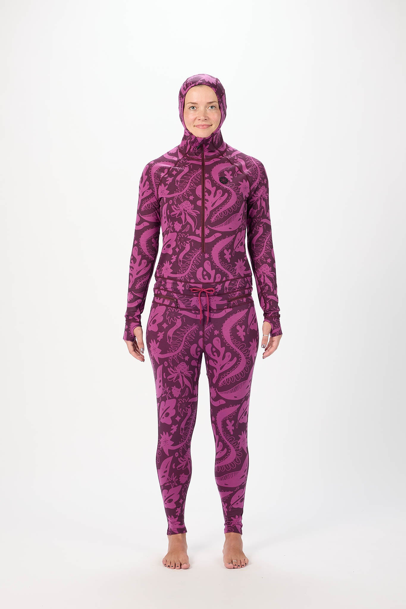 A woman modeling a full-body purple and black paisley-patterned onesie  with a matching hood, standing against a plain white studio background.