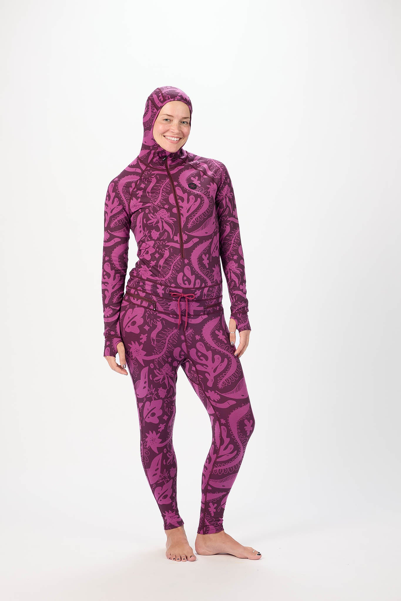 A woman modeling a full-body purple and black paisley-patterned onesie  with a matching hood, standing against a plain white studio background.