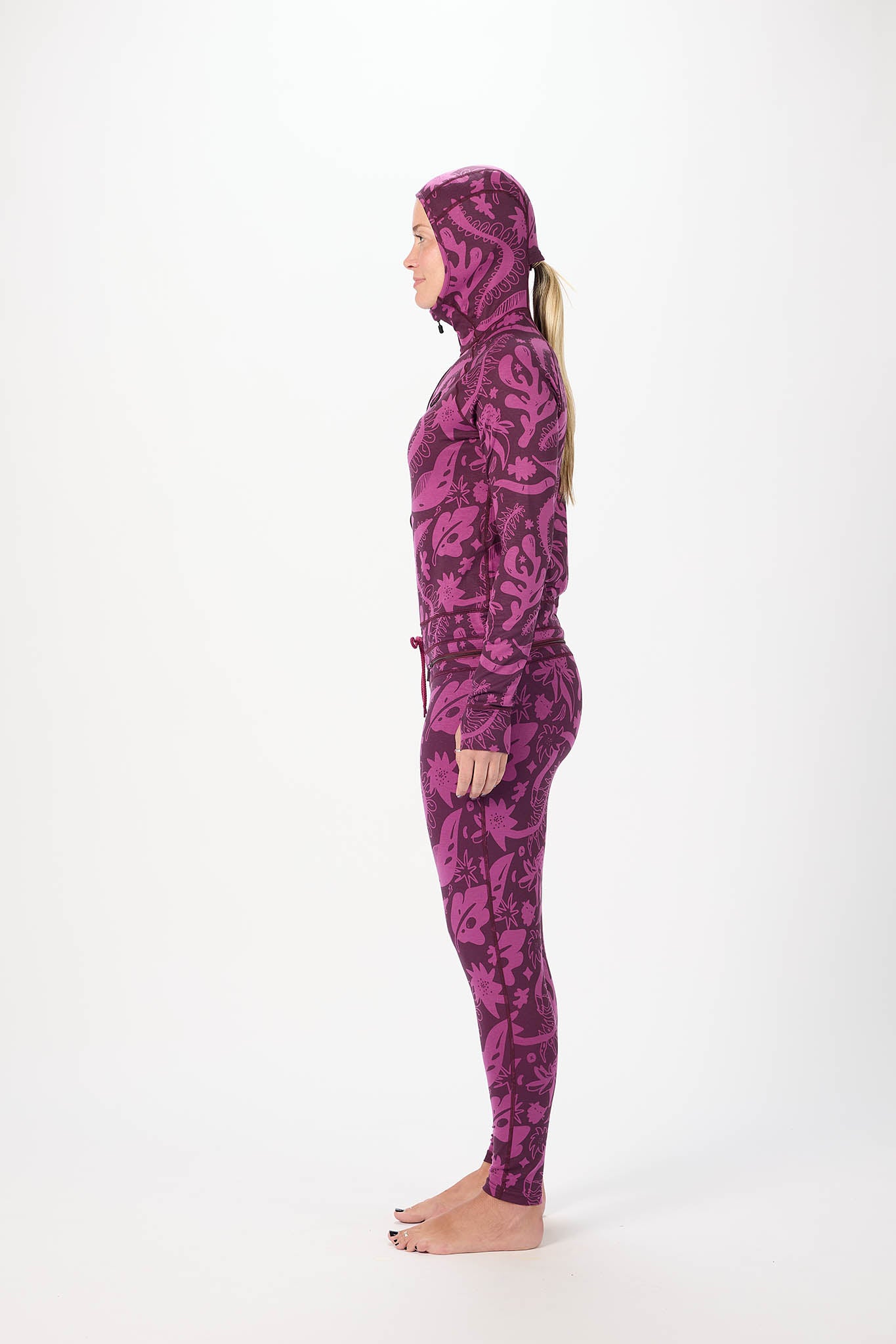 inside profile A woman modeling a full-body purple and black paisley-patterned onesie  with a matching hood, standing against a plain white studio background.