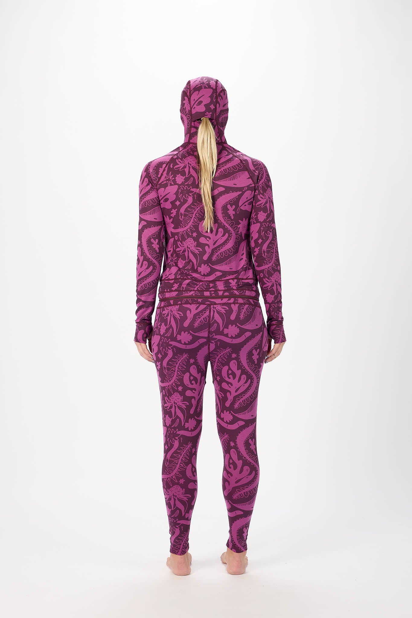 A woman viewed from behind wearing a matching hooded long-sleeve top and leggings set with an abstract swirling pattern in magenta and purple tones, standing against a plain light gray studio background.