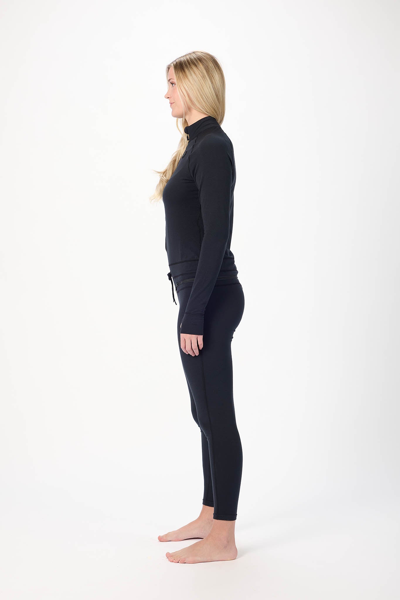 A woman standing in profile against a plain white studio background, wearing a black long-sleeve mock-neck top and matching black leggings.