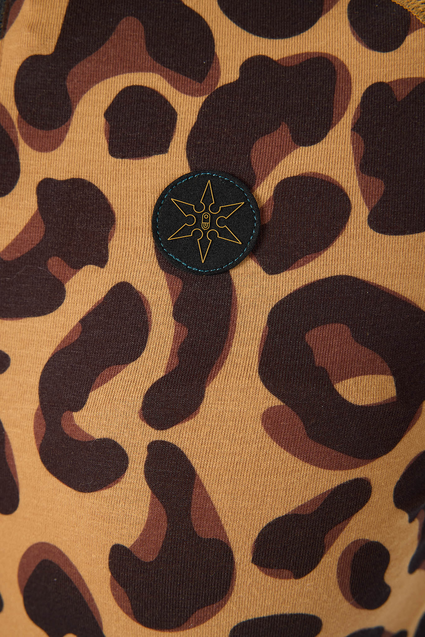 Close-up of a leopard print garment featuring a tan base with dark brown and black spots, and a small circular black patch with a gold star logo near the center.