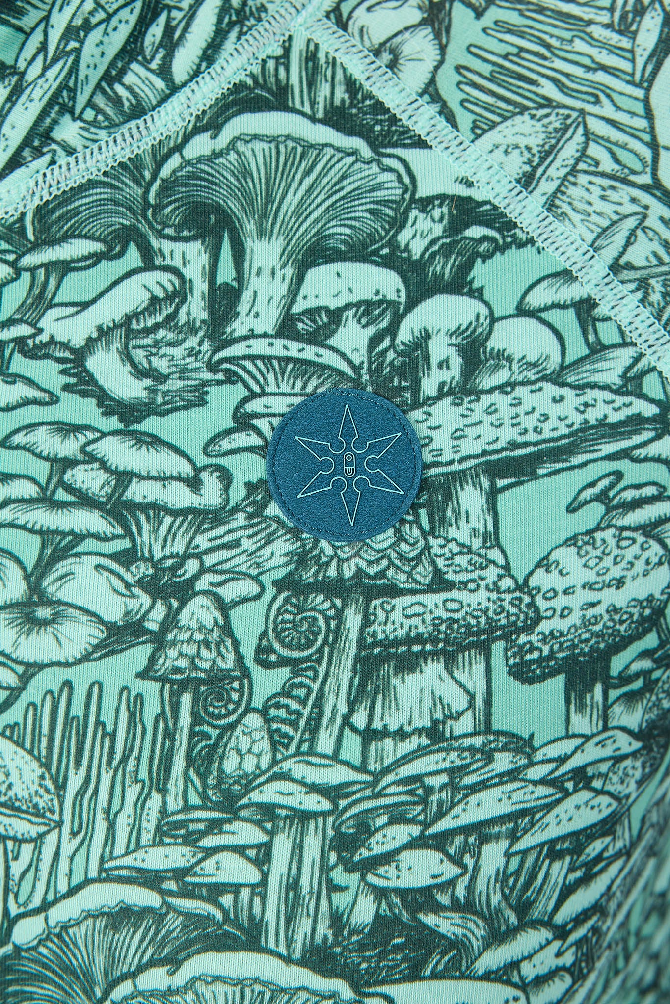 Close-up of a fabric surface featuring a detailed black line-art illustration of mushrooms and forest plants in a mint green and teal colorway, with a circular blue patch containing a white star design near the center.