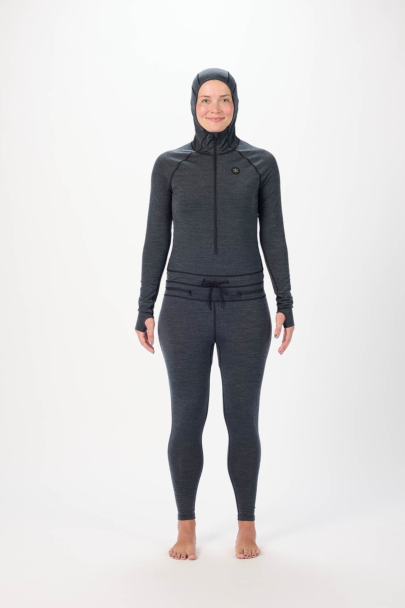 A woman wearing a black full-body athletic suit with a high-neck zip-front top, matching pants, and a hood, standing against a plain white studio background.