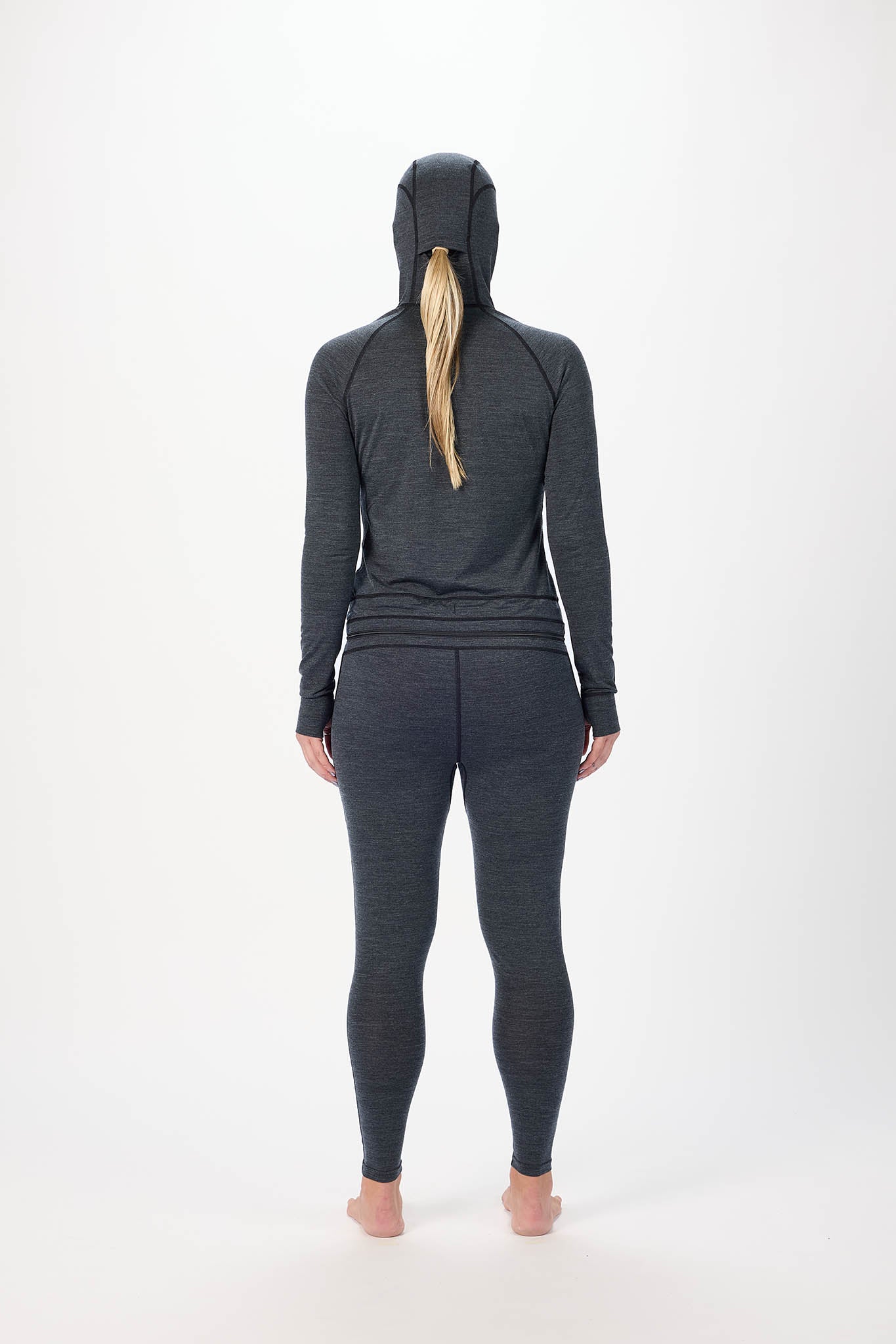 A woman viewed from the back wearing a dark gray heathered hooded long-sleeve top and matching leggings, standing barefoot against a plain white studio background.