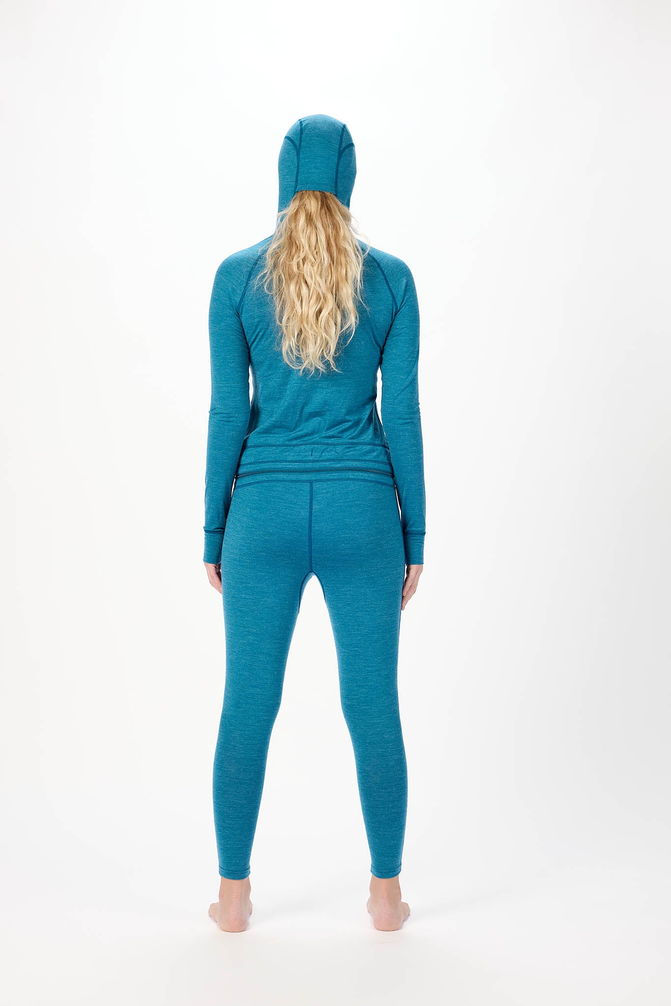 A woman viewed from behind wearing a matching teal blue three-piece activewear set consisting of a long-sleeve top, leggings, and a hooded head covering, against a plain white studio background.