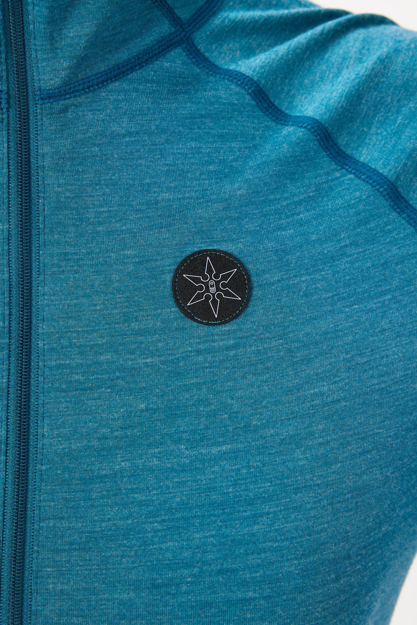 Close-up of a teal heathered athletic jacket with a black circular chest patch featuring a white stylized compass or star logo.