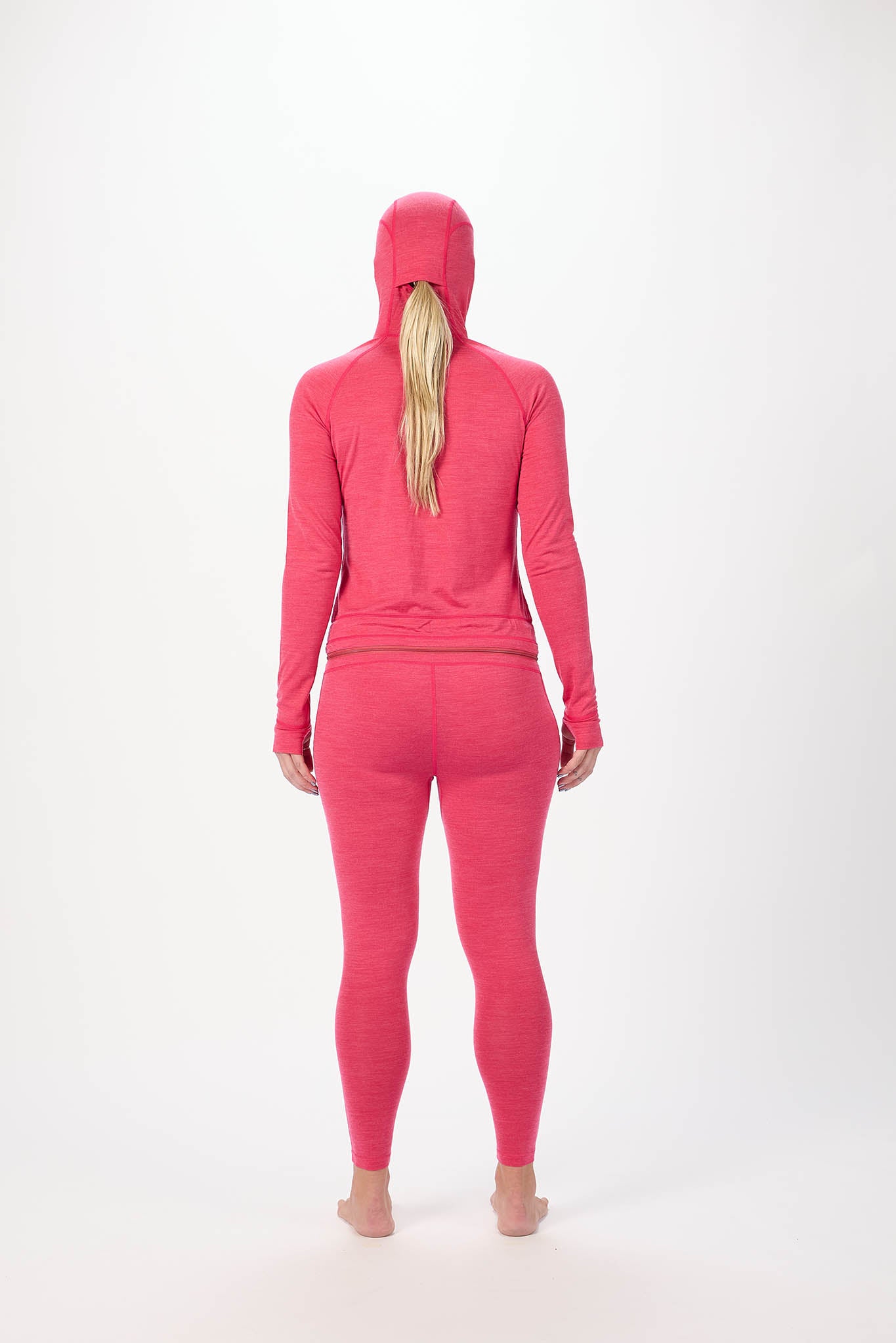 A woman viewed from behind wearing a bright pink matching activewear set consisting of a hooded long-sleeve top and full-length leggings, standing against a plain white studio background.