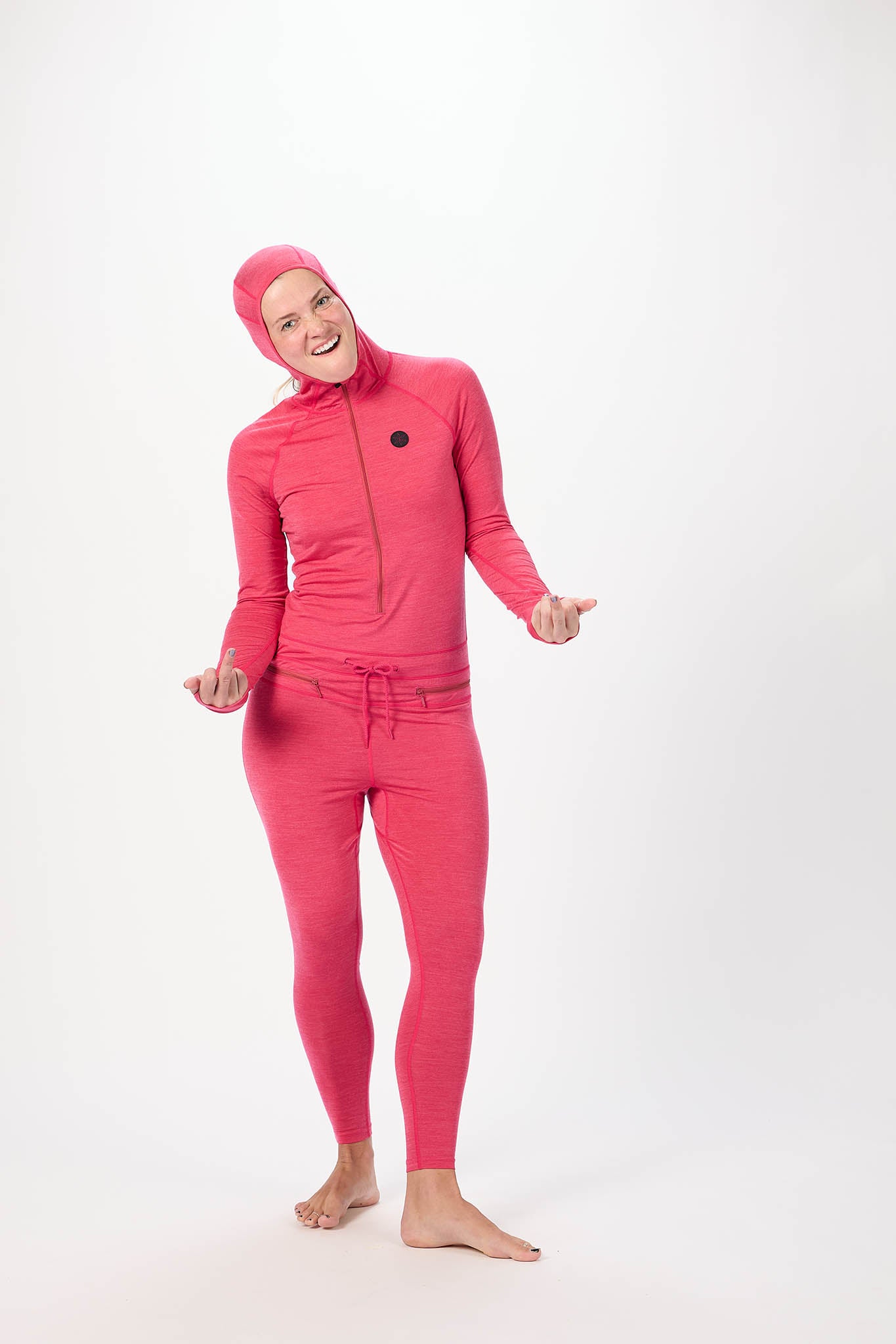 A person wearing a bright pink full-body hooded suit with a front zipper, matching pants, and a drawstring waist, standing barefoot against a plain light gray studio background.