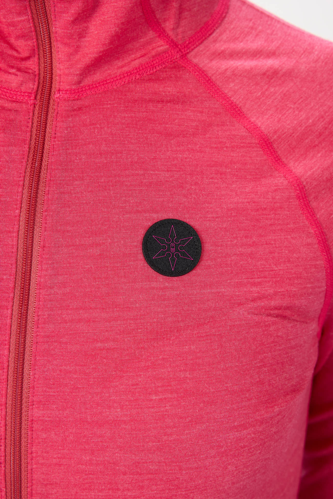 Close-up of a heathered pink athletic zip-up jacket with a black circular chest patch featuring a pink geometric star design.