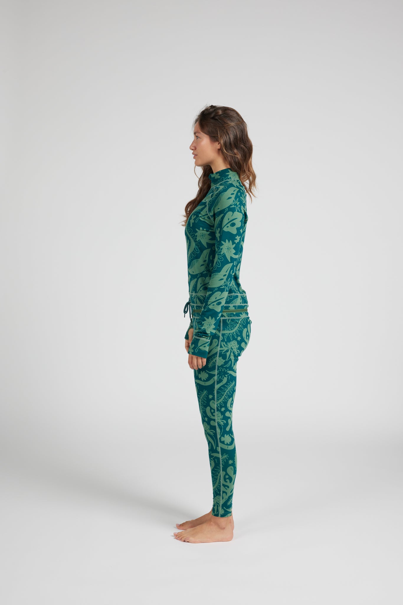 A woman stands in profile wearing a matching two-piece activewear set with a green and teal tropical leaf pattern, photographed against a neutral light gray studio background.