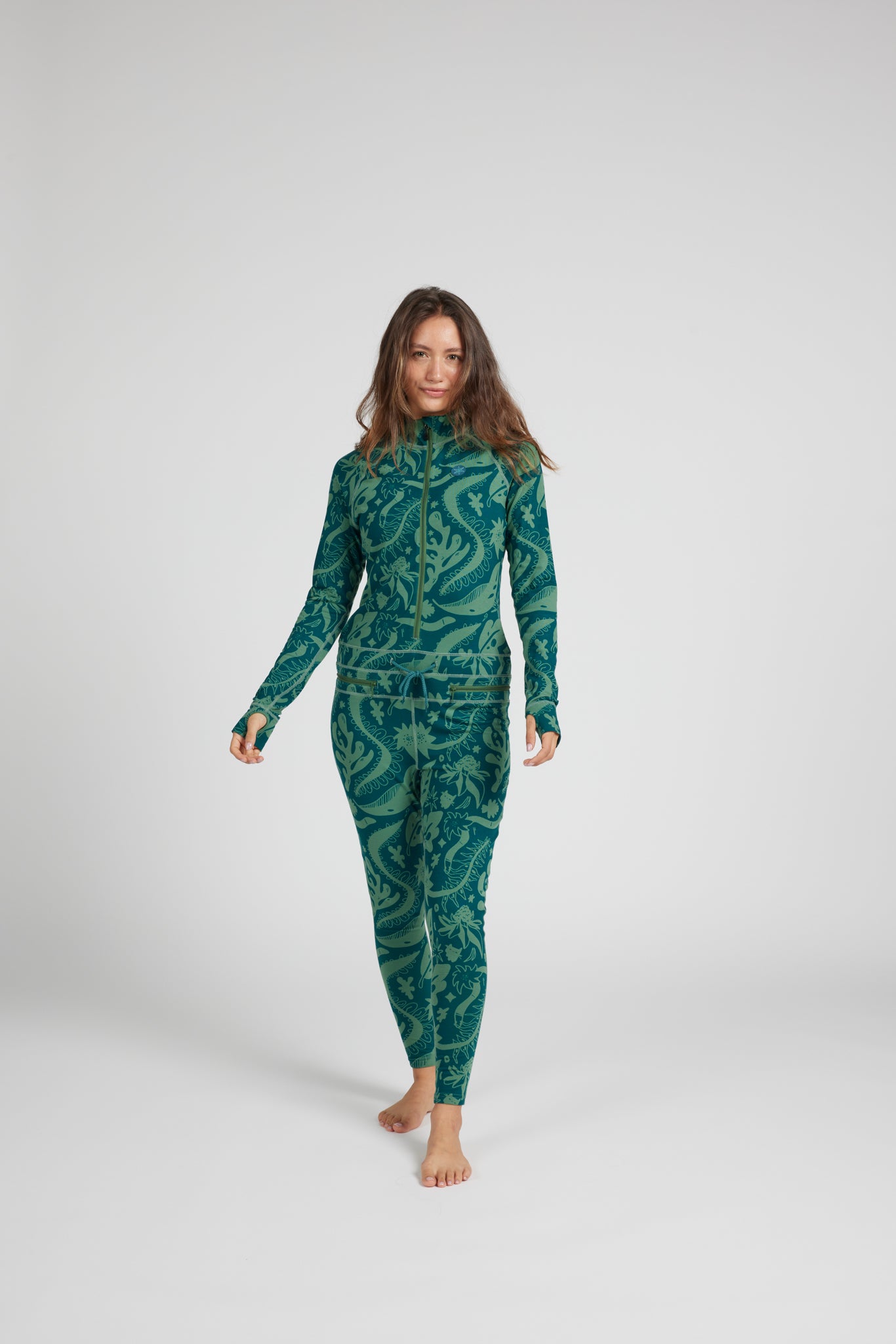 A woman modeling a full-body long-sleeve wetsuit with a green and teal abstract pattern, standing against a light gray studio background.
