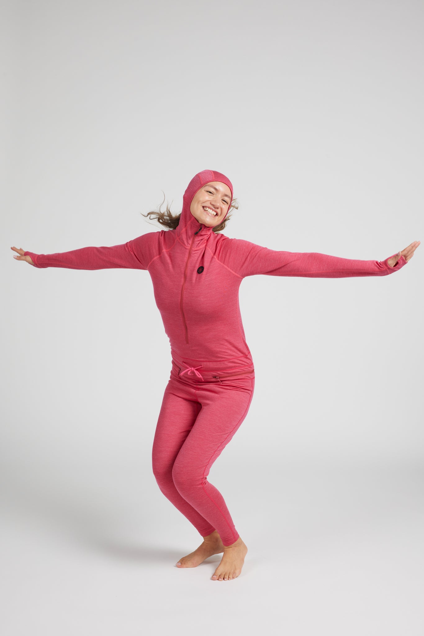 A woman wearing a bright pink full-body hooded suit with long sleeves and leggings, posing with her arms outstretched against a light gray studio background.