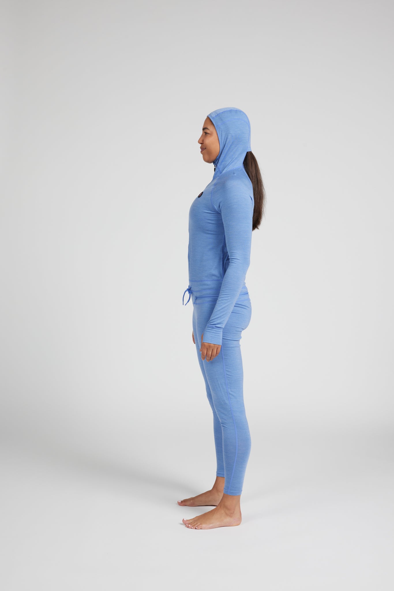 A woman stands in profile wearing a light blue full-body hooded knit outfit against a plain light gray studio background.