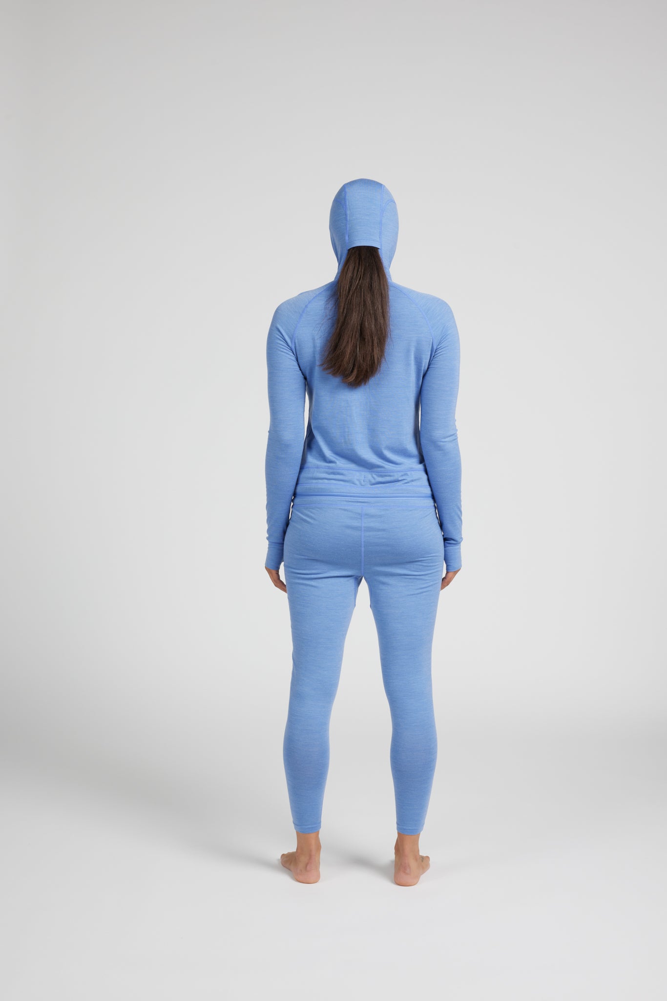 A woman viewed from behind wearing a light blue one-piece long-sleeve hooded outfit, standing barefoot against a plain light gray studio background.