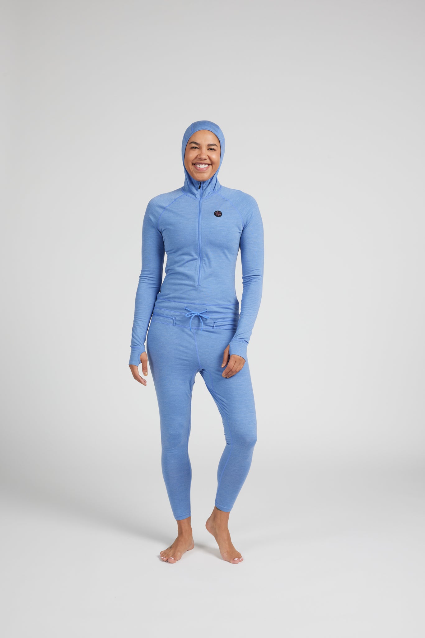 A woman modeling a light blue full-body hooded suit with a high-zip top and matching leggings against a plain light gray studio background.