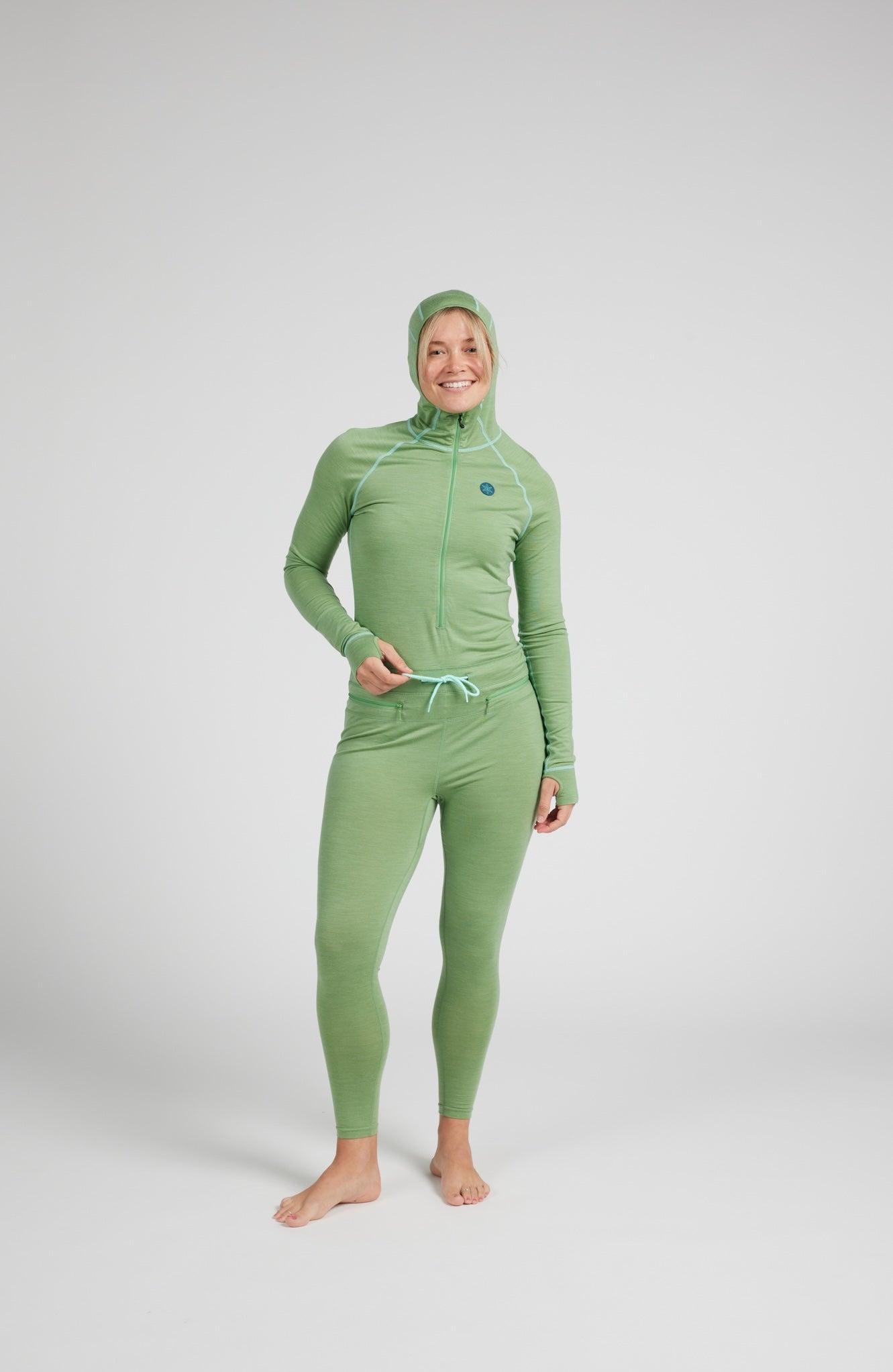 A woman modeling a light green full-body wetsuit with a hood, long sleeves, and a drawstring waist, standing against a light gray studio background.