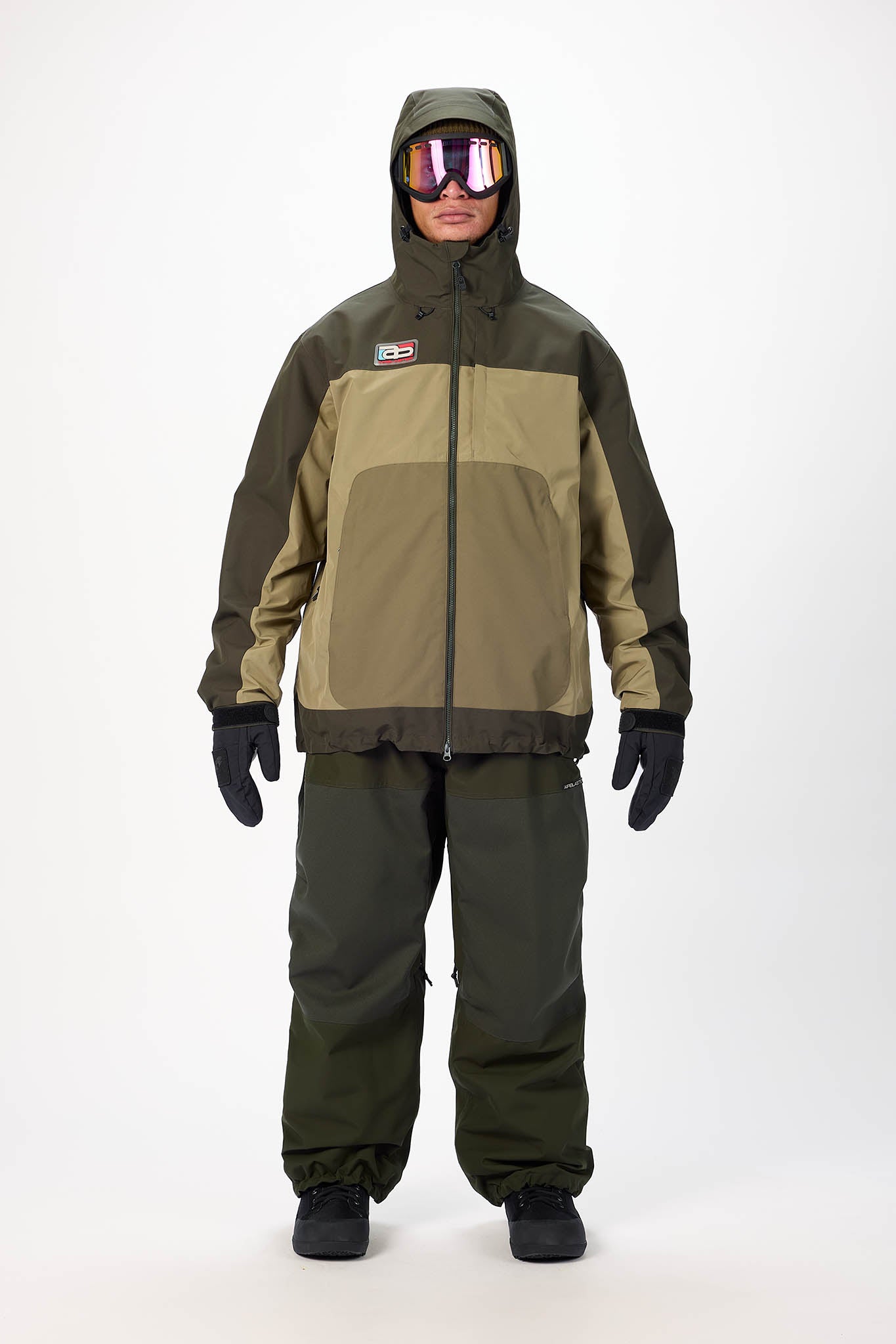 Person wearing a green and brown snowsuit with goggles on a white background