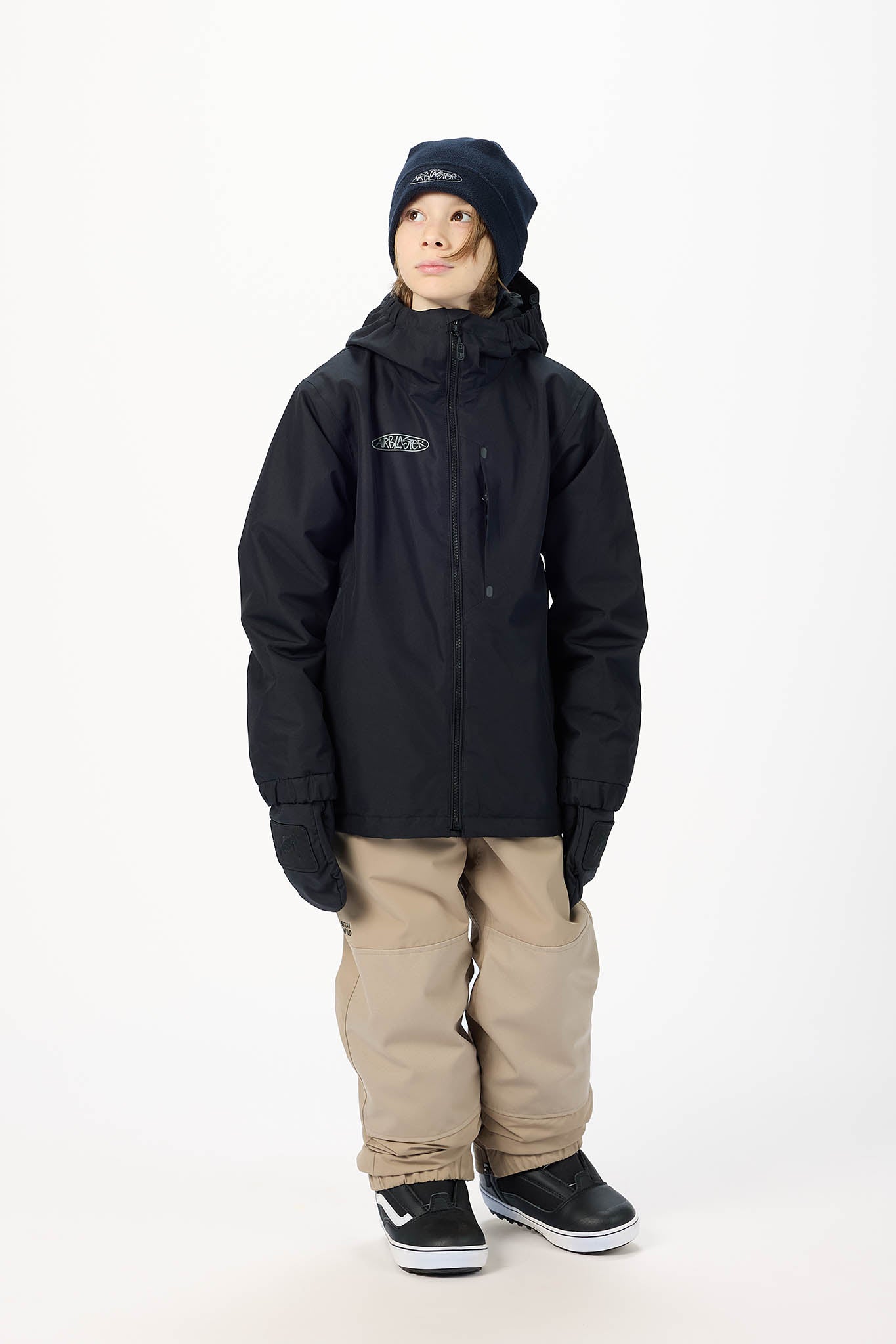 A child model wearing a black hooded snowboard jacket, tan snow pants, black gloves, and a black beanie, standing against a plain white studio background.