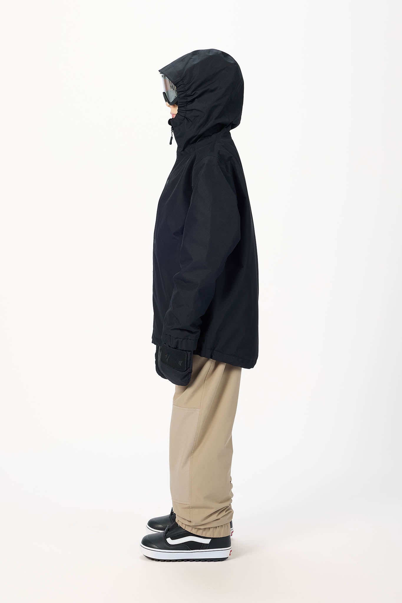 A person shown in profile wearing a black hooded jacket, beige pants, and black sneakers against a plain light gray studio background.
