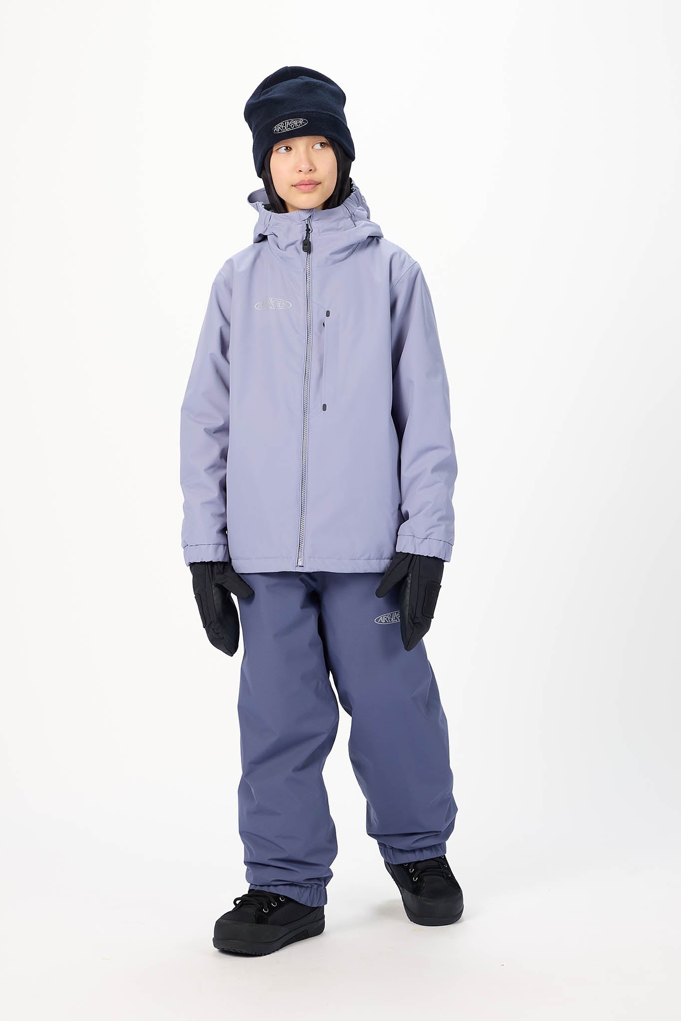 A child modeling a light purple ski jacket, matching purple snow pants, black gloves, and a dark navy beanie against a plain white studio background.