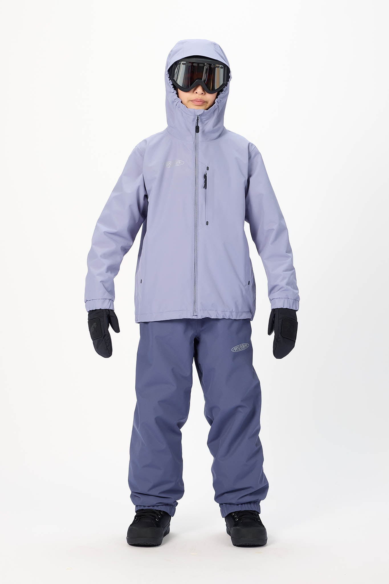 A child model wearing a light lavender ski jacket, matching snow pants, black gloves, and goggles, standing against a plain white studio background.