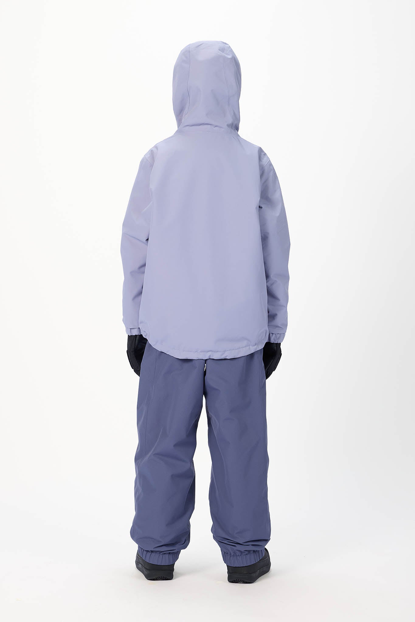 A child standing with their back to the camera, wearing a light lavender hooded jacket and slate blue snow pants against a plain white studio background.
