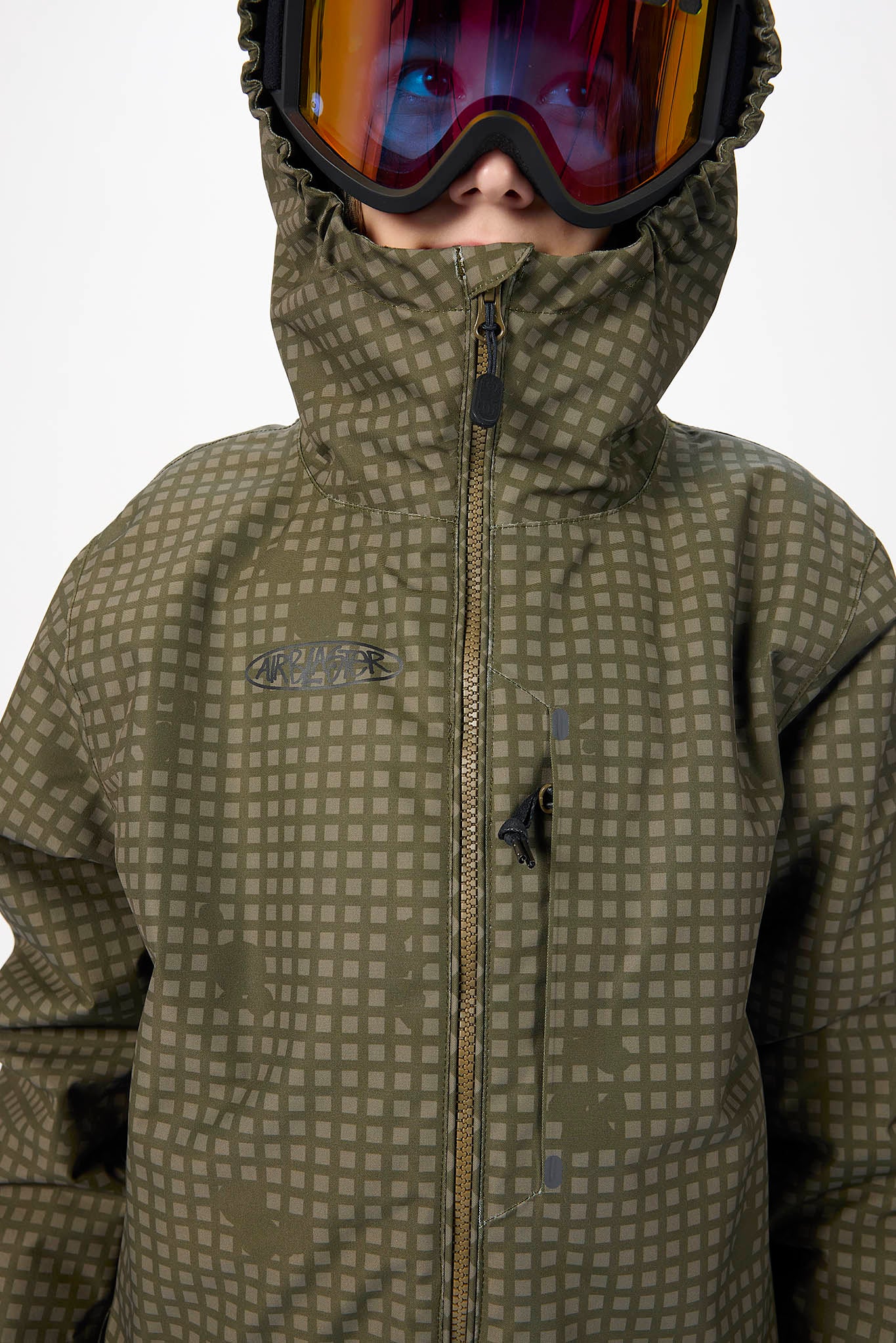 A close-up of a child wearing an olive green ski jacket with a small grid pattern, a high hood, and ski goggles with a reflective orange lens.