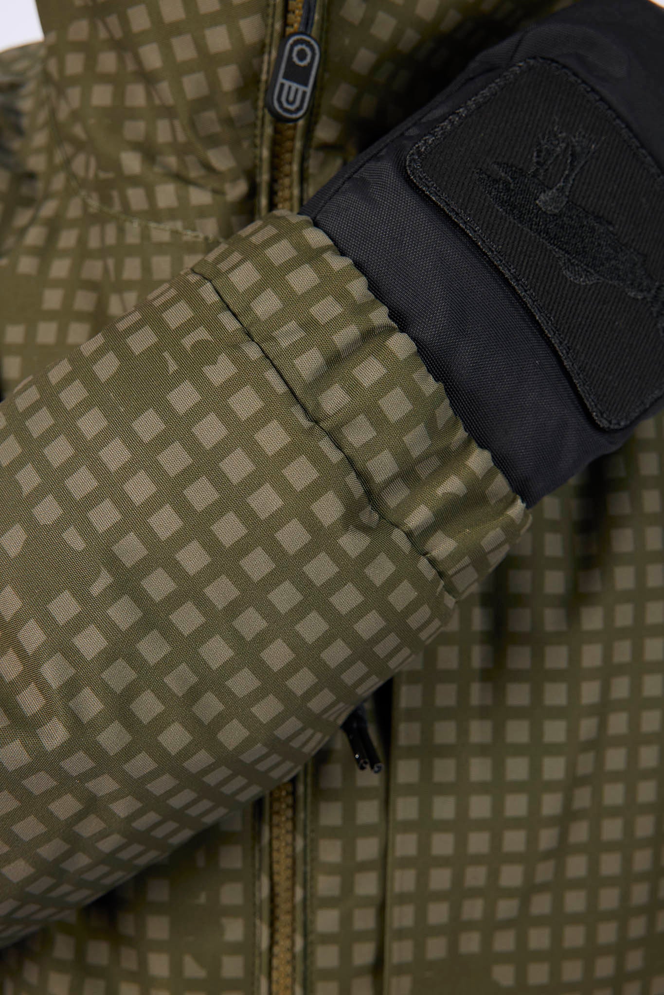 Close-up of a puffer jacket sleeve with a beige and olive green checkered pattern, a black cuff, and a black patch featuring a stylized logo.