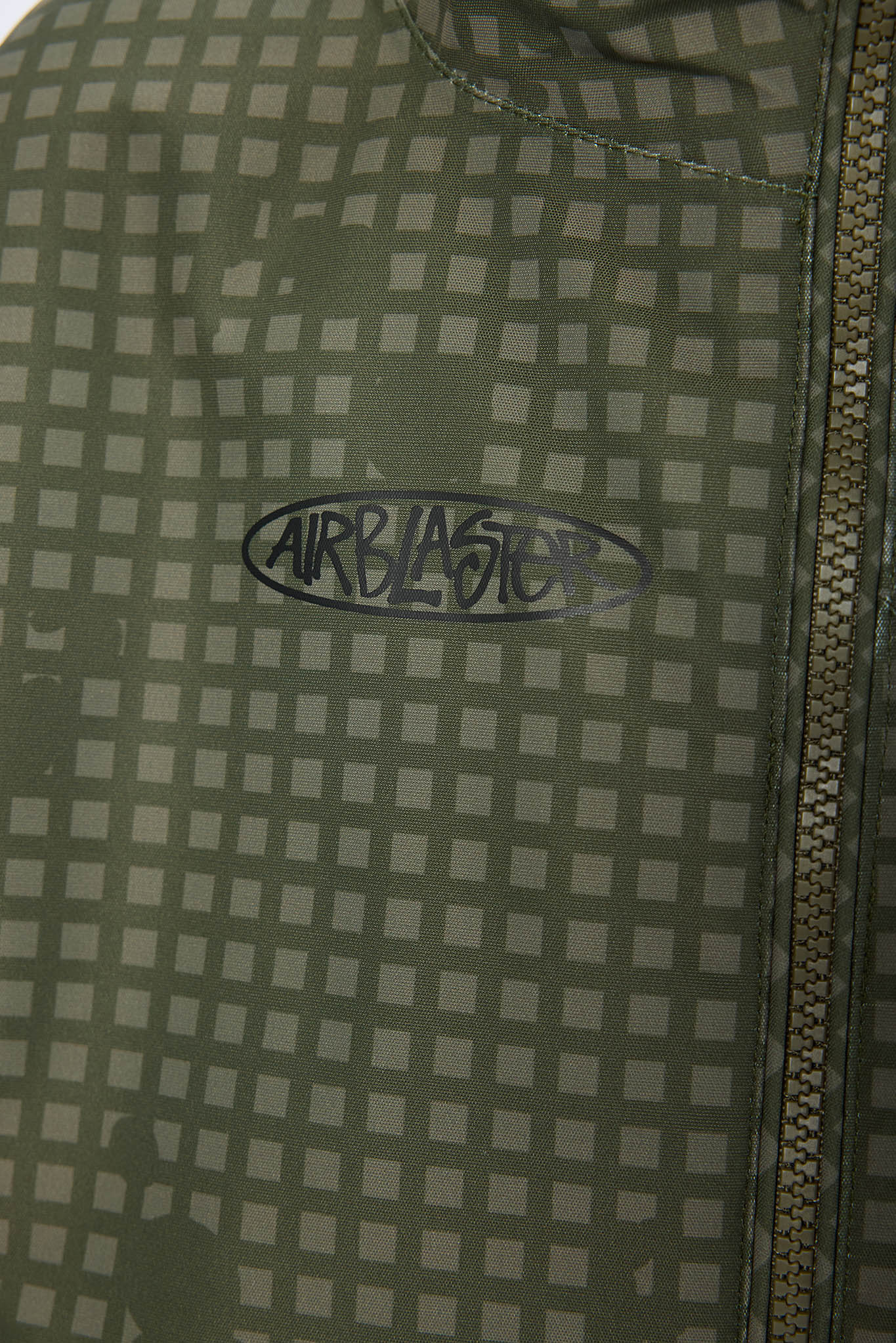 Close-up of a green and gray checkered patterned fabric with a black Airblaster logo in the center and a zipper on the right side.